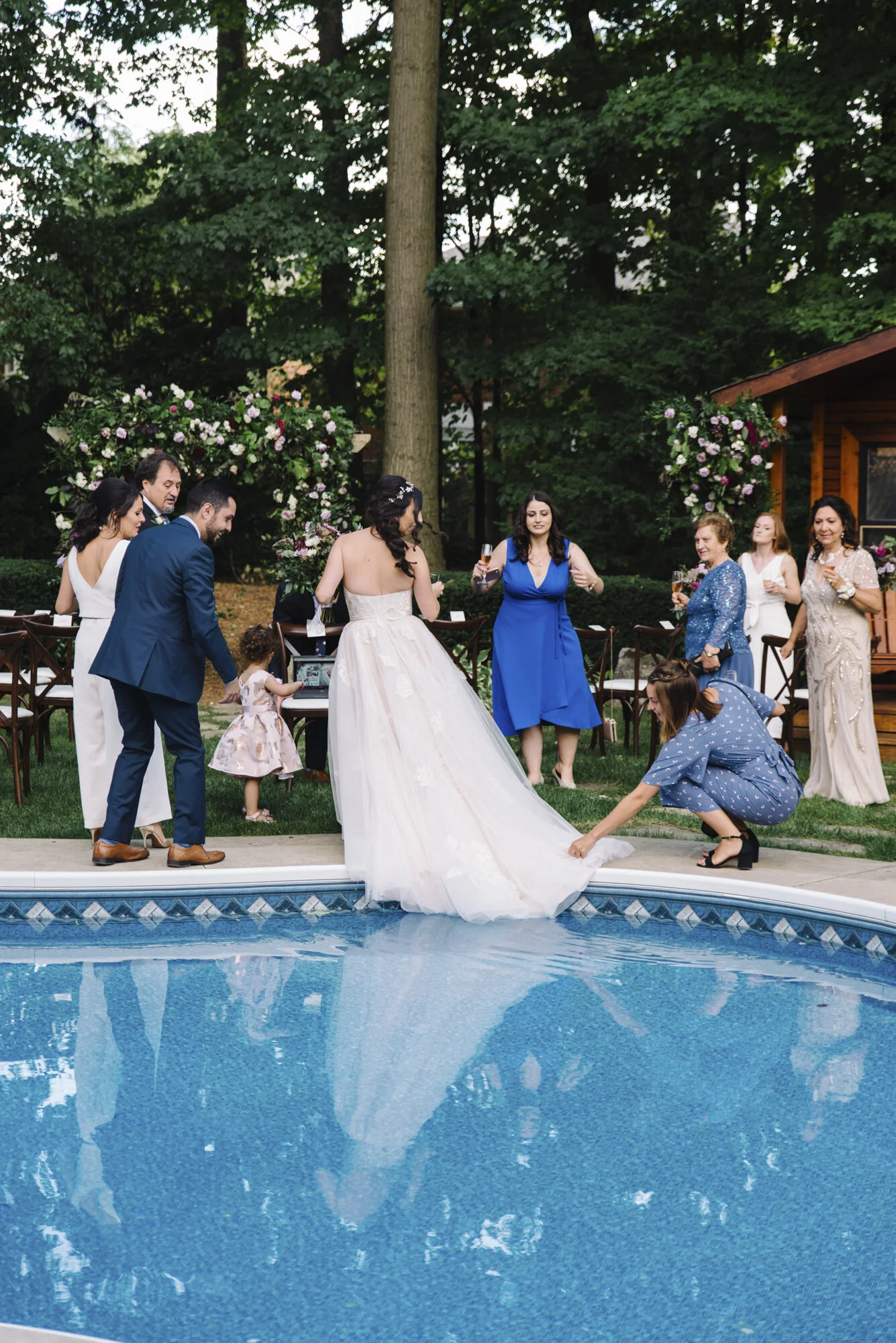 Candid photo of a bride's wedding dress dipping into the pool as guests react around her — she is completely unbothered