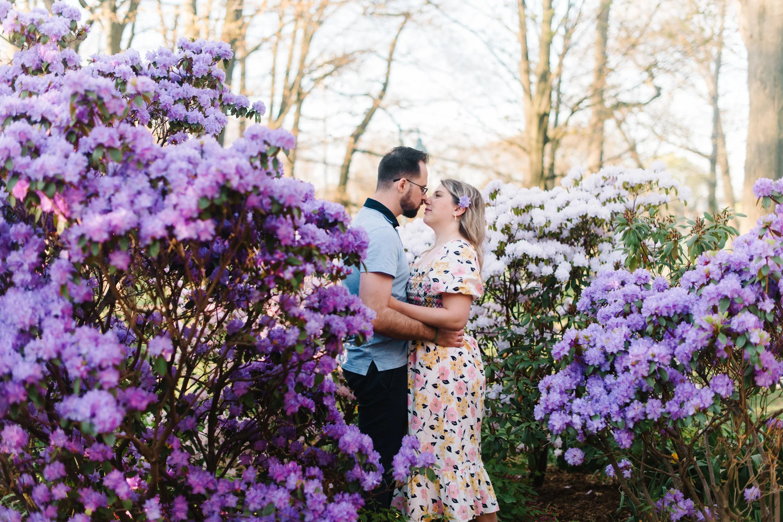 Romantic portrait of a couple standing in a vibrant lilac bush facing each other with the sunrise behind them at Woodbine Beach, Toronto