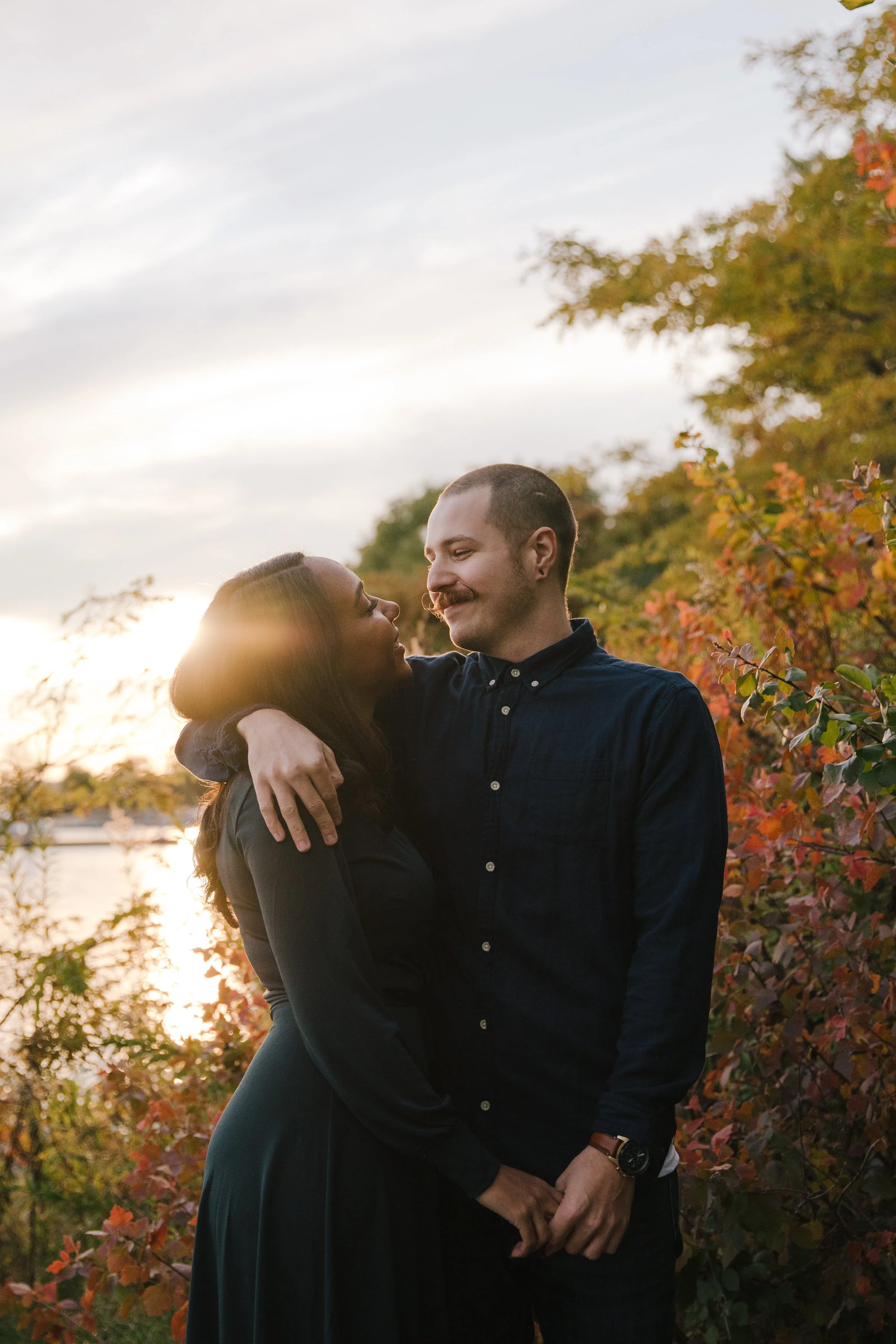 Romantic engagement photo of a couple embracing at sunset surrounded by vibrant fall colours in Toronto, sun peeking through the trees