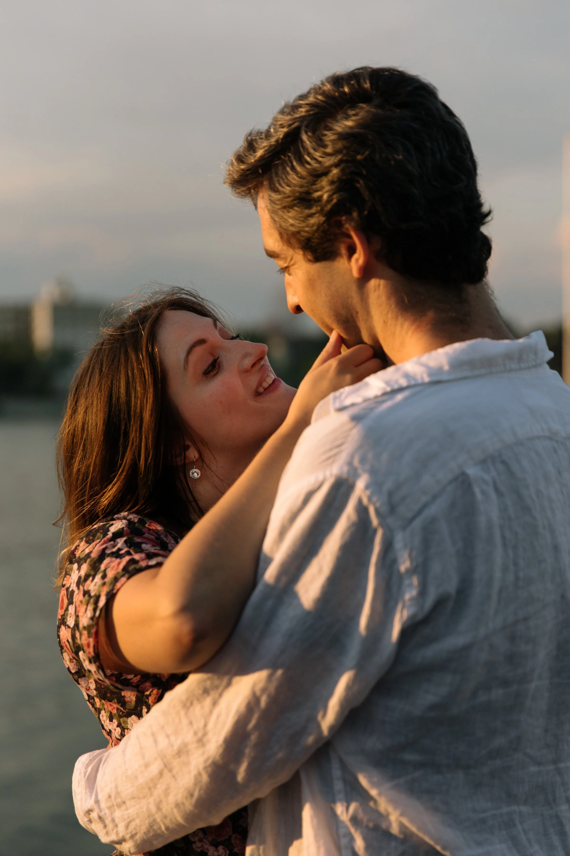 Candid engagement portrait of a couple standing by the water at sunset, one partner looking lovingly at the other