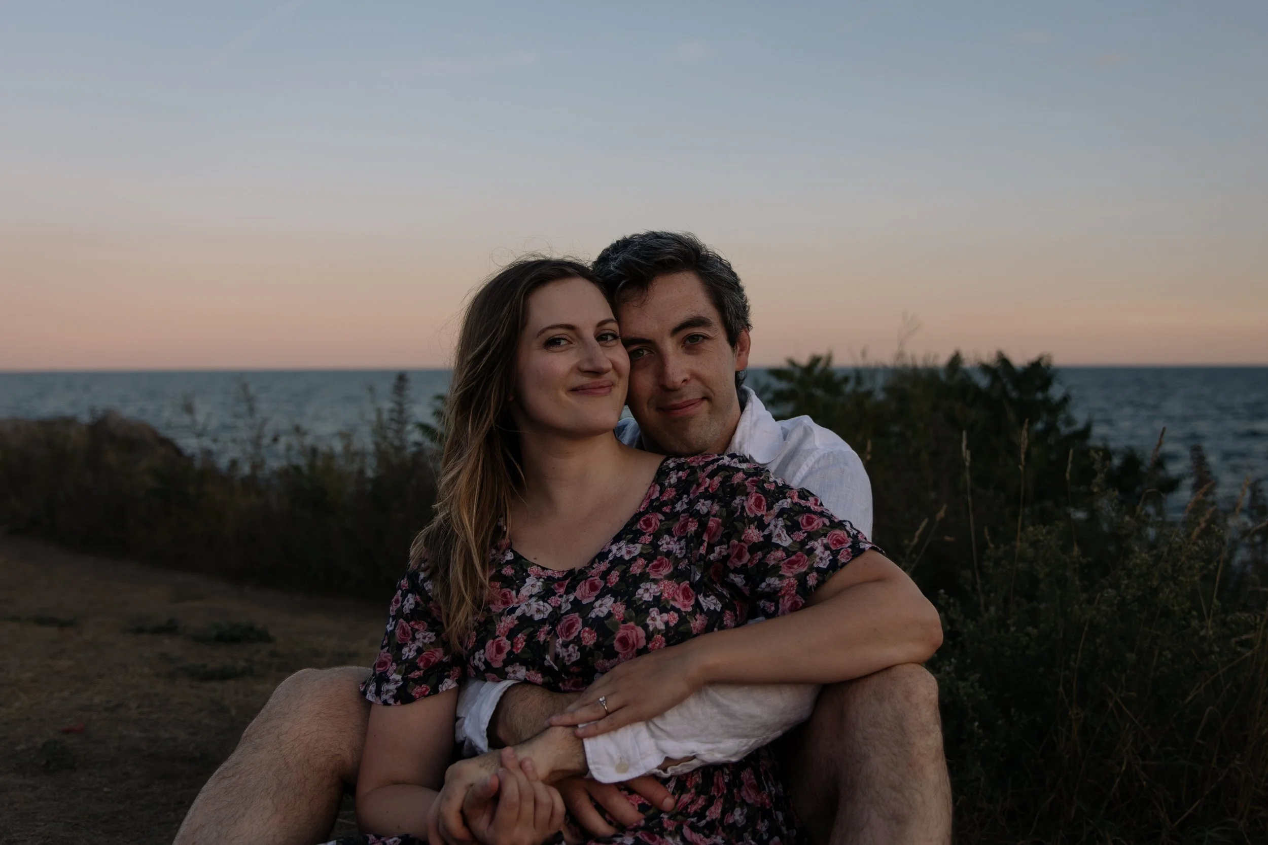 Engaged couple sitting and holding each other as the sun sets over the Toronto waterfront