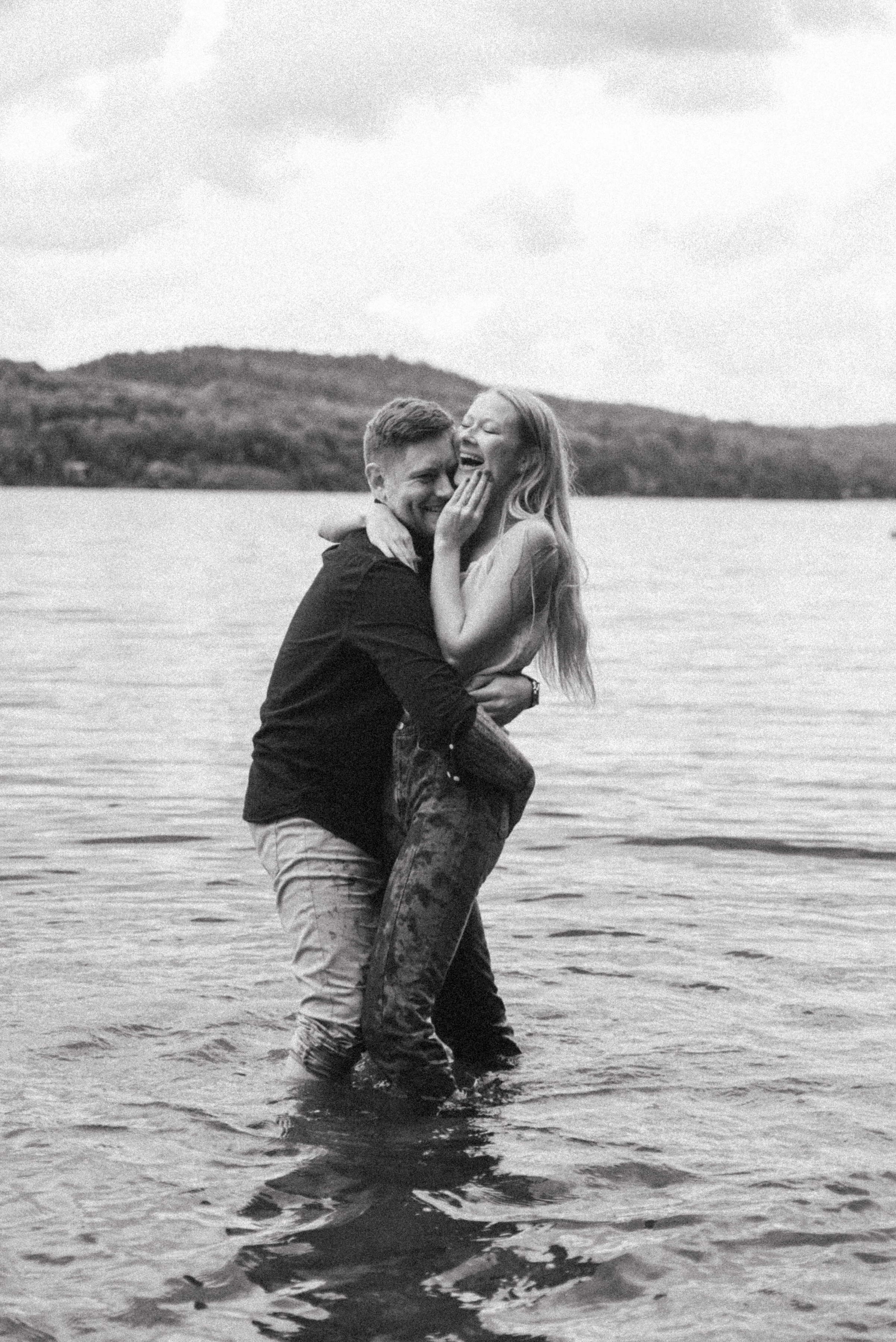 Black and white film photo of a couple playfully embracing in the water, smiling and laughing during their engagement session in Muskoka