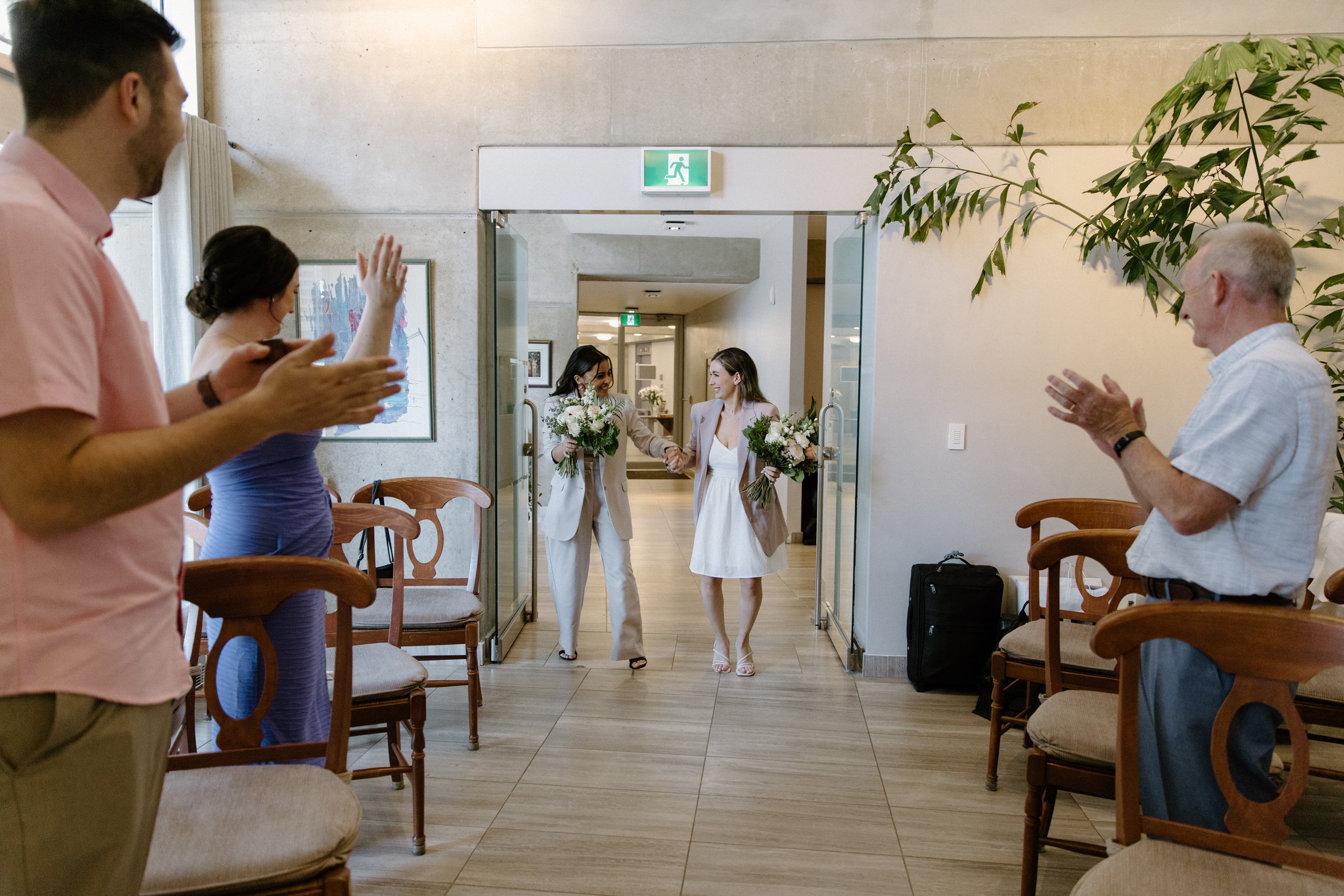 Candid photo of two brides entering their wedding ceremony at Toronto City Hall as guests stand and cheer