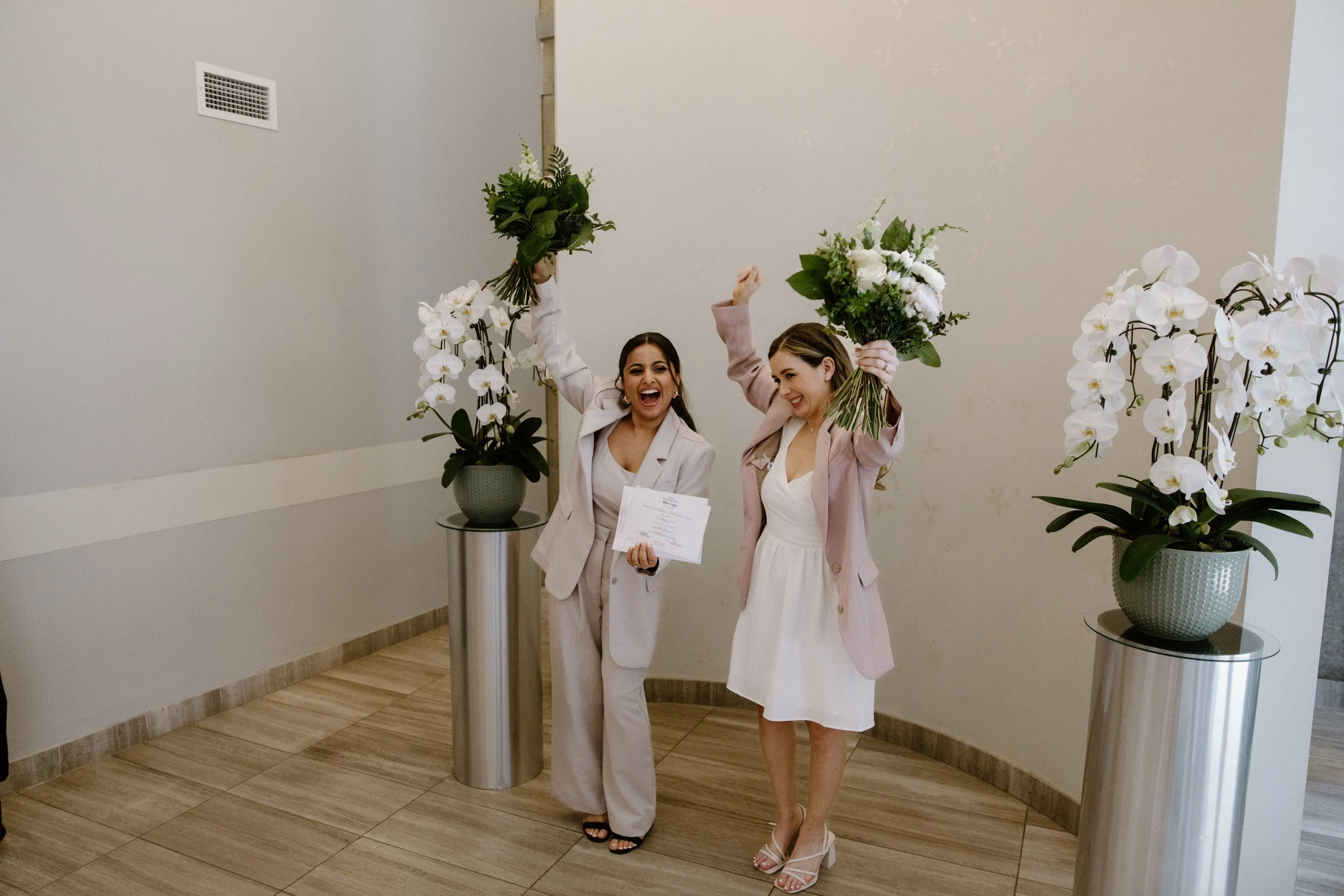 Candid photo of two brides joyfully holding their marriage certificate after their same-sex elopement at Toronto City Hall