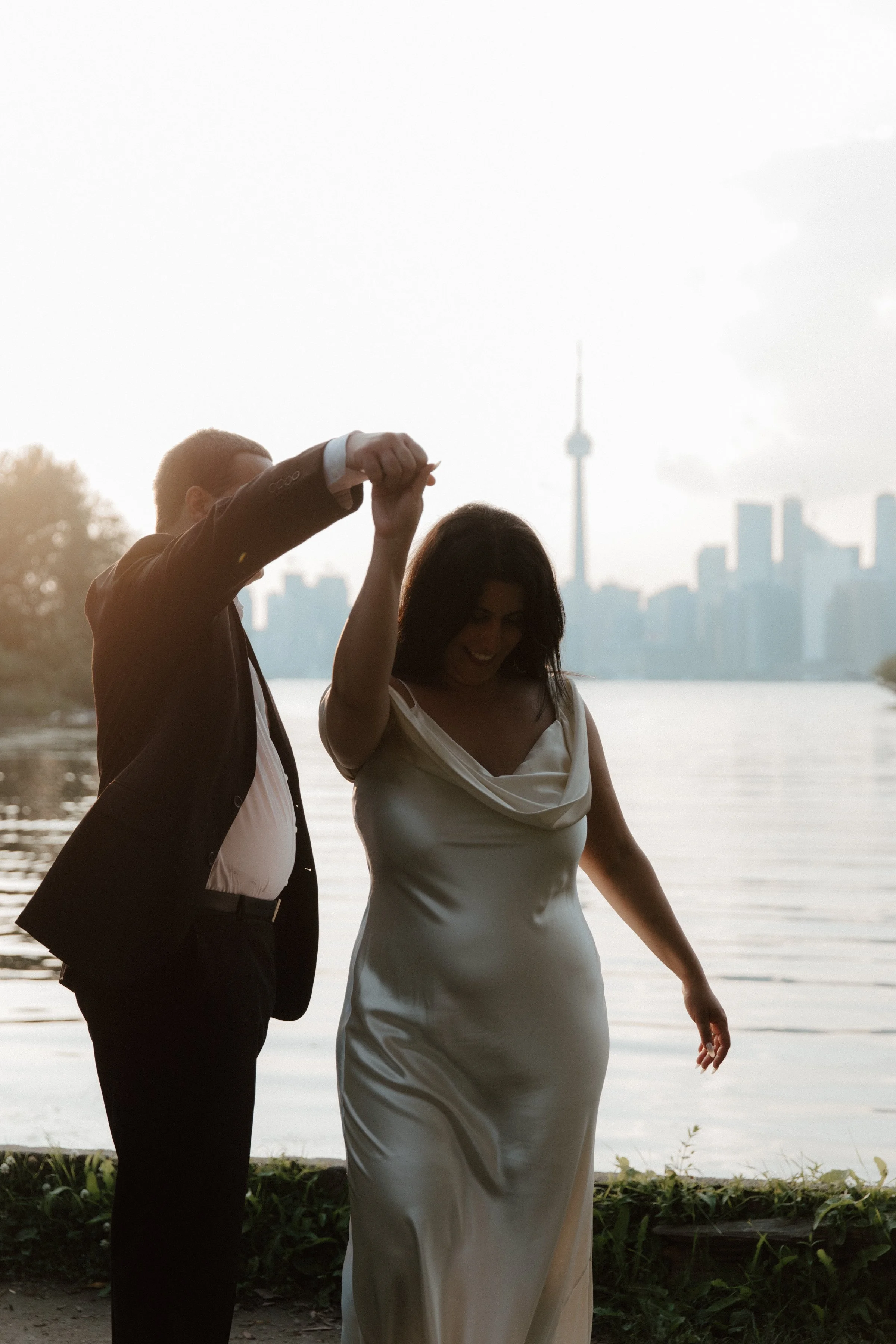 A couple wearing wedding attire dance's at sunset in front of the Toronto skyline