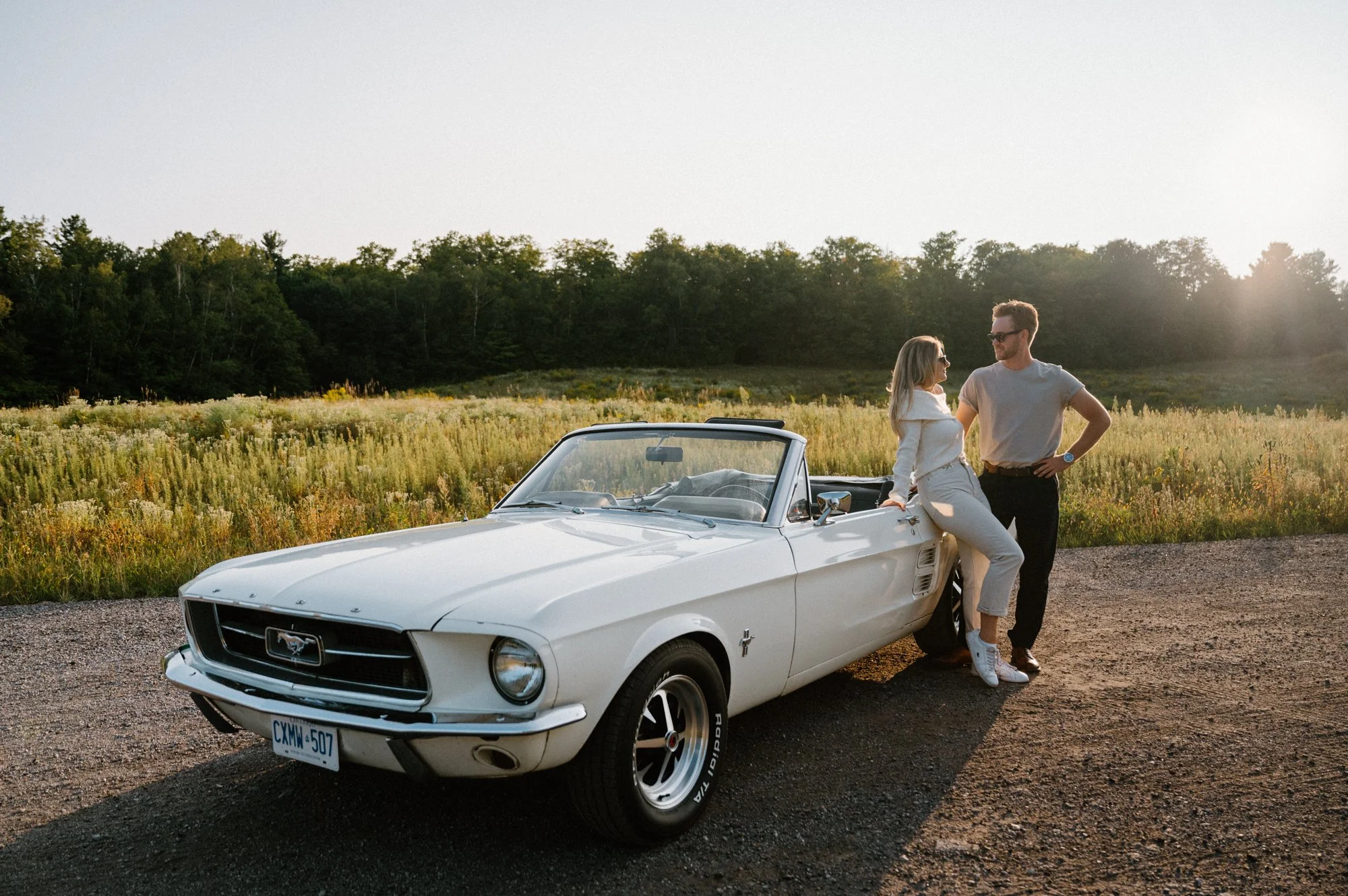 Editorial engagement portrait of a couple sitting on the hood of a vintage Mustang at golden hour in the Kawarthas
