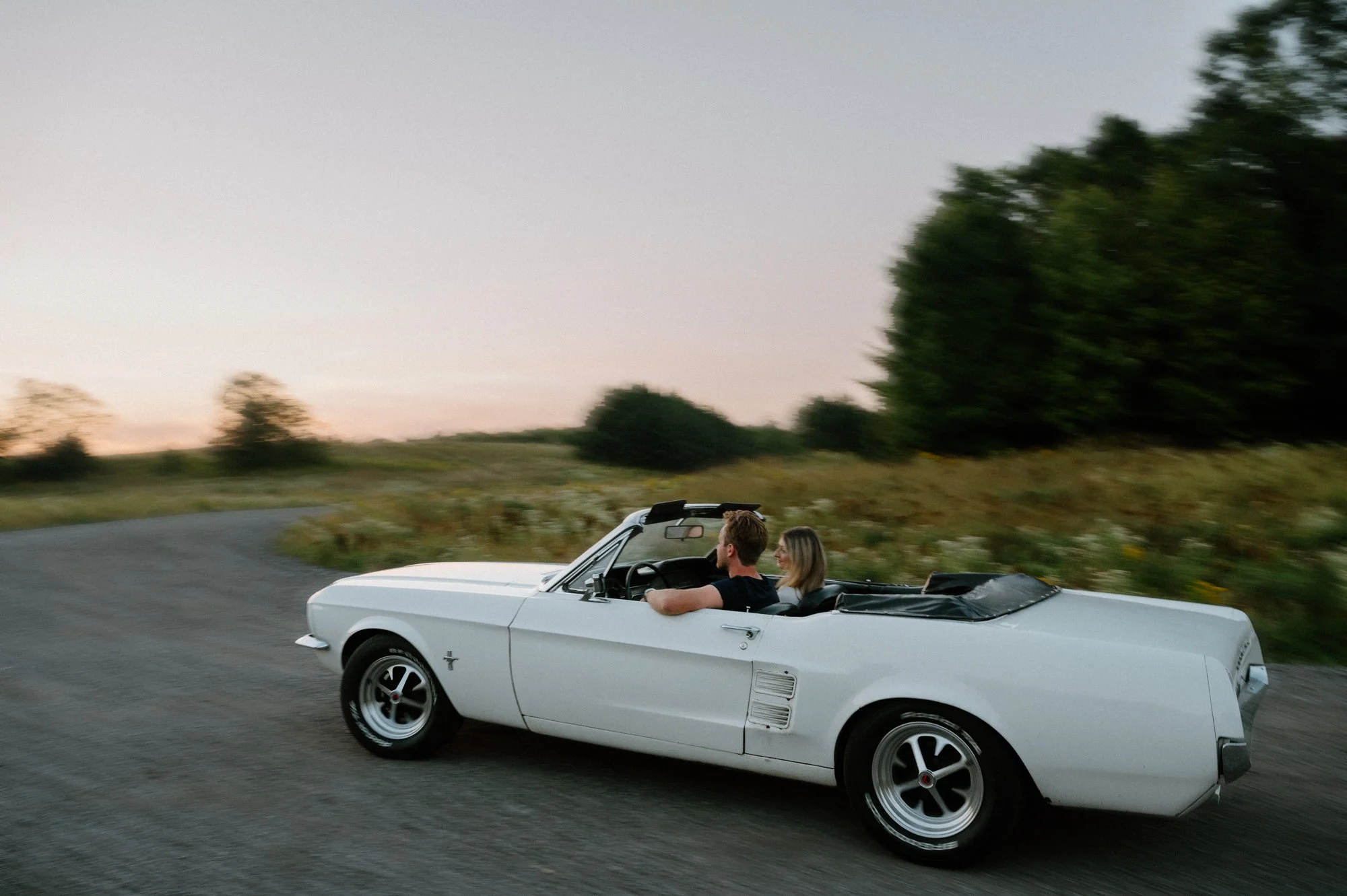 Engaged couple driving away in a vintage Mustang at sunset during their engagement session in the Kawarthas