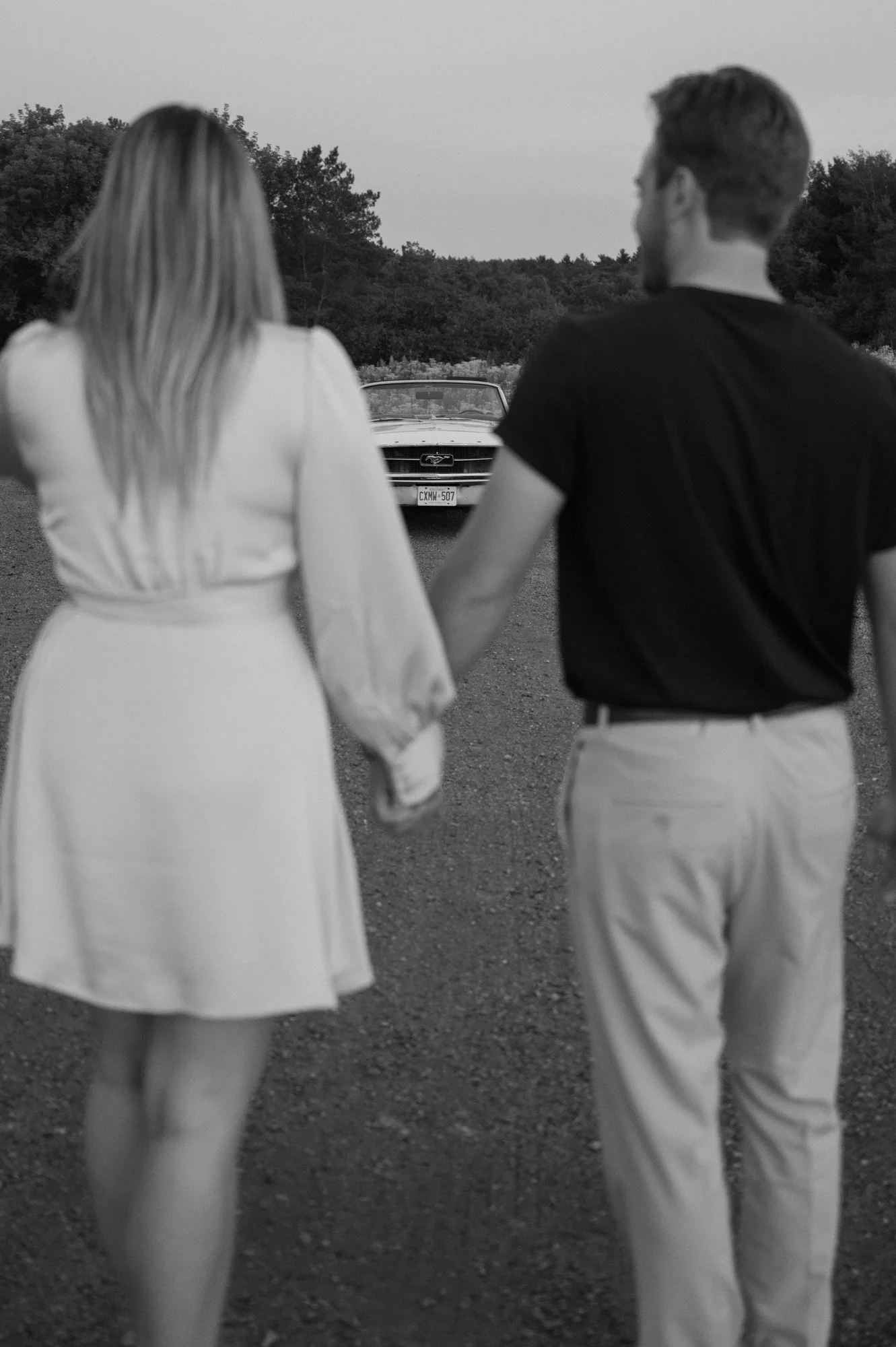 Engaged couple walking towards a vintage Mustang in an open field, car in sharp focus during their Ontario engagement session