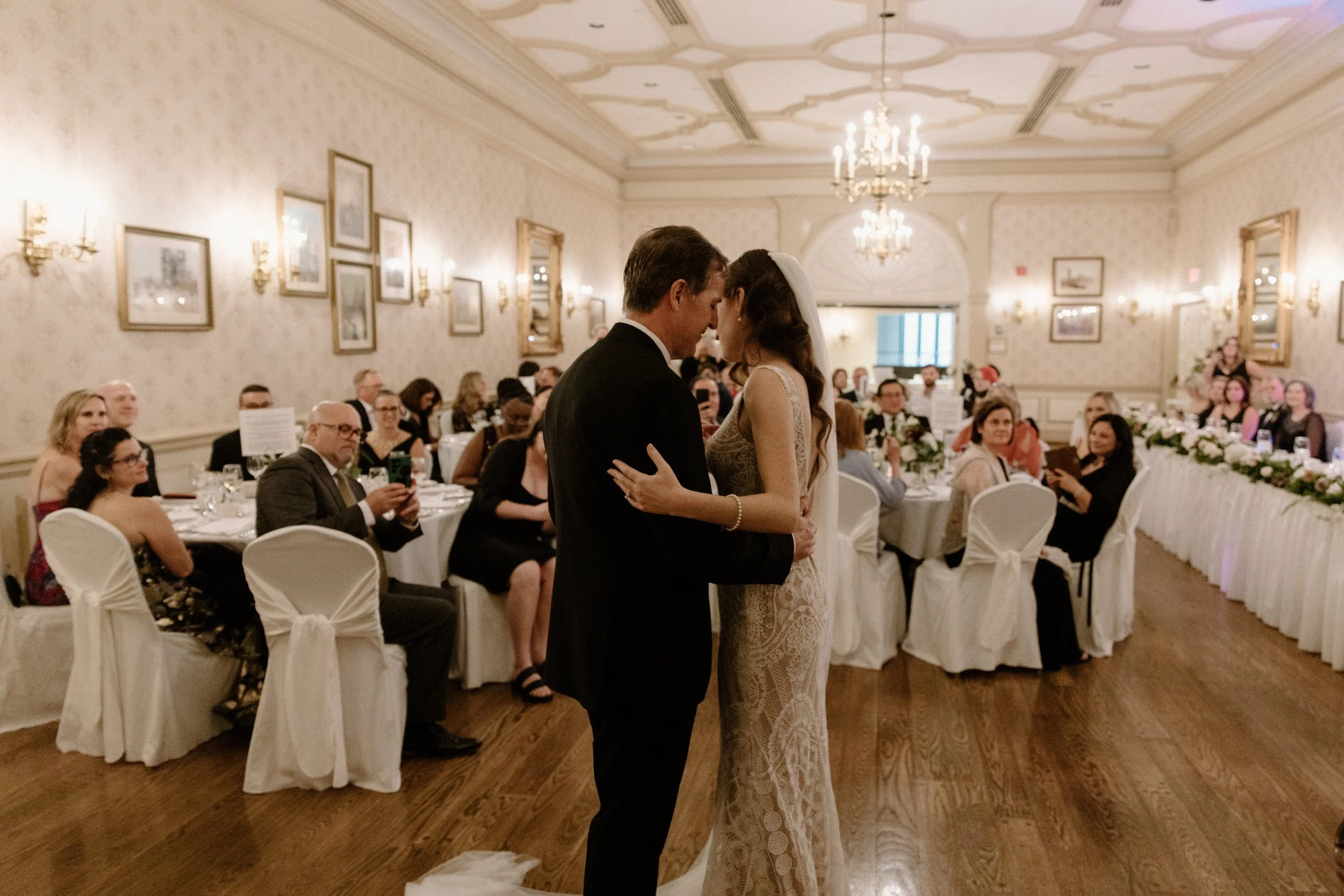 Wide photo of a bride and groom sharing their first dance with smiling guests watching behind them at their Toronto wedding reception