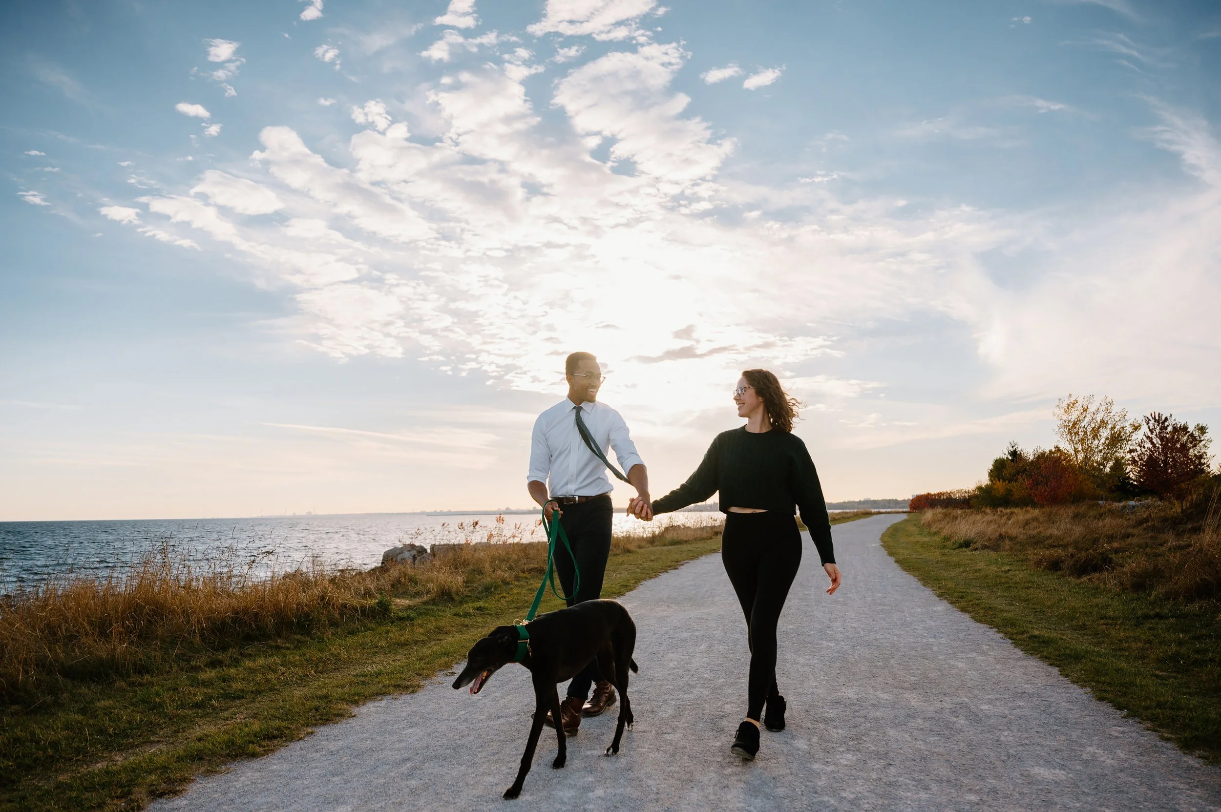 Candid photo of an engaged couple walking their dog along the waterfront at golden hour during their engagement session
