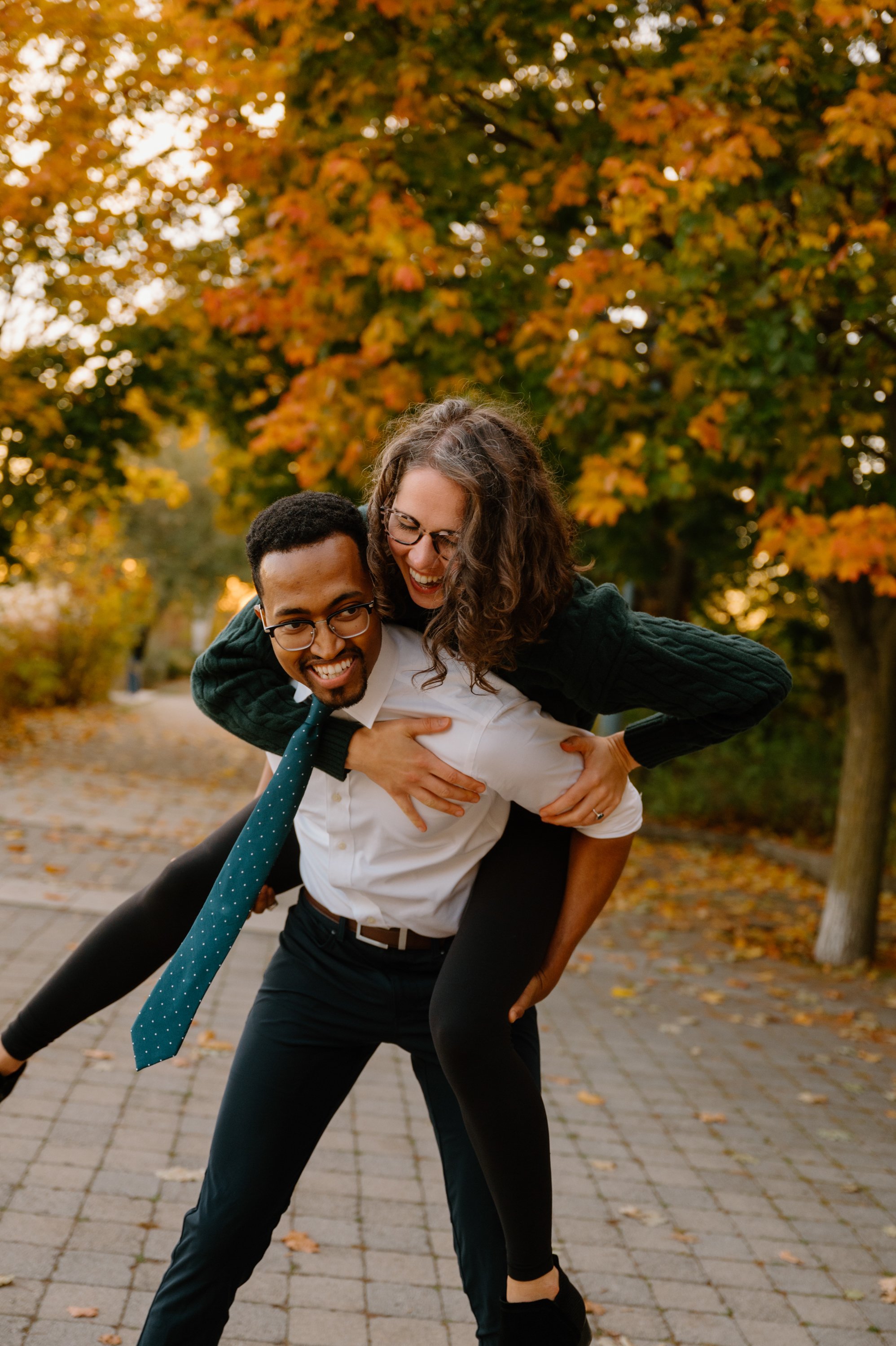 Candid and playful photo of an engaged couple laughing together with fall colours and a warm sunset in the background