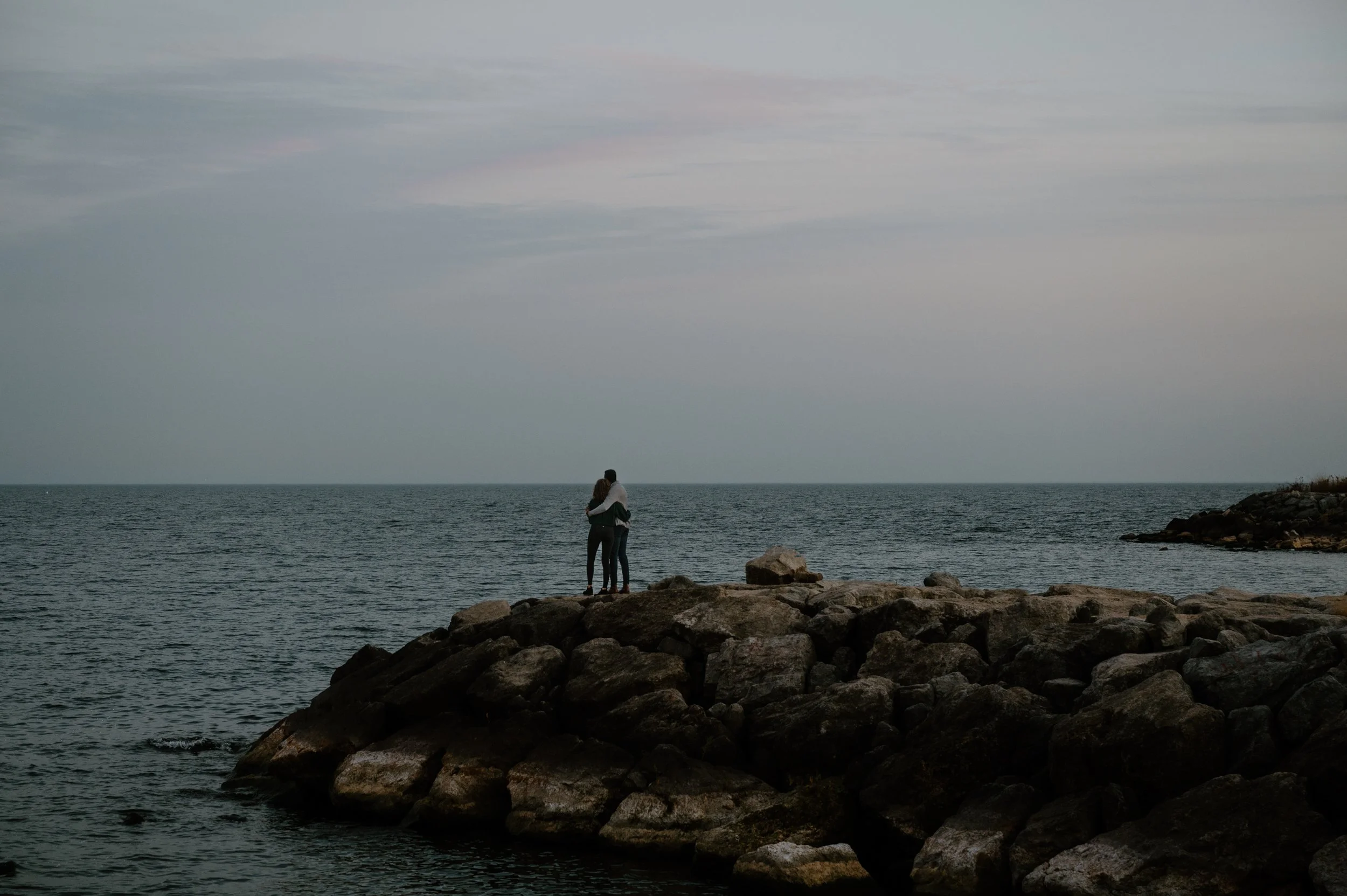 Wide documentary landscape photo of an engaged couple looking out over the water, reminiscent of a Vancouver coastal setting