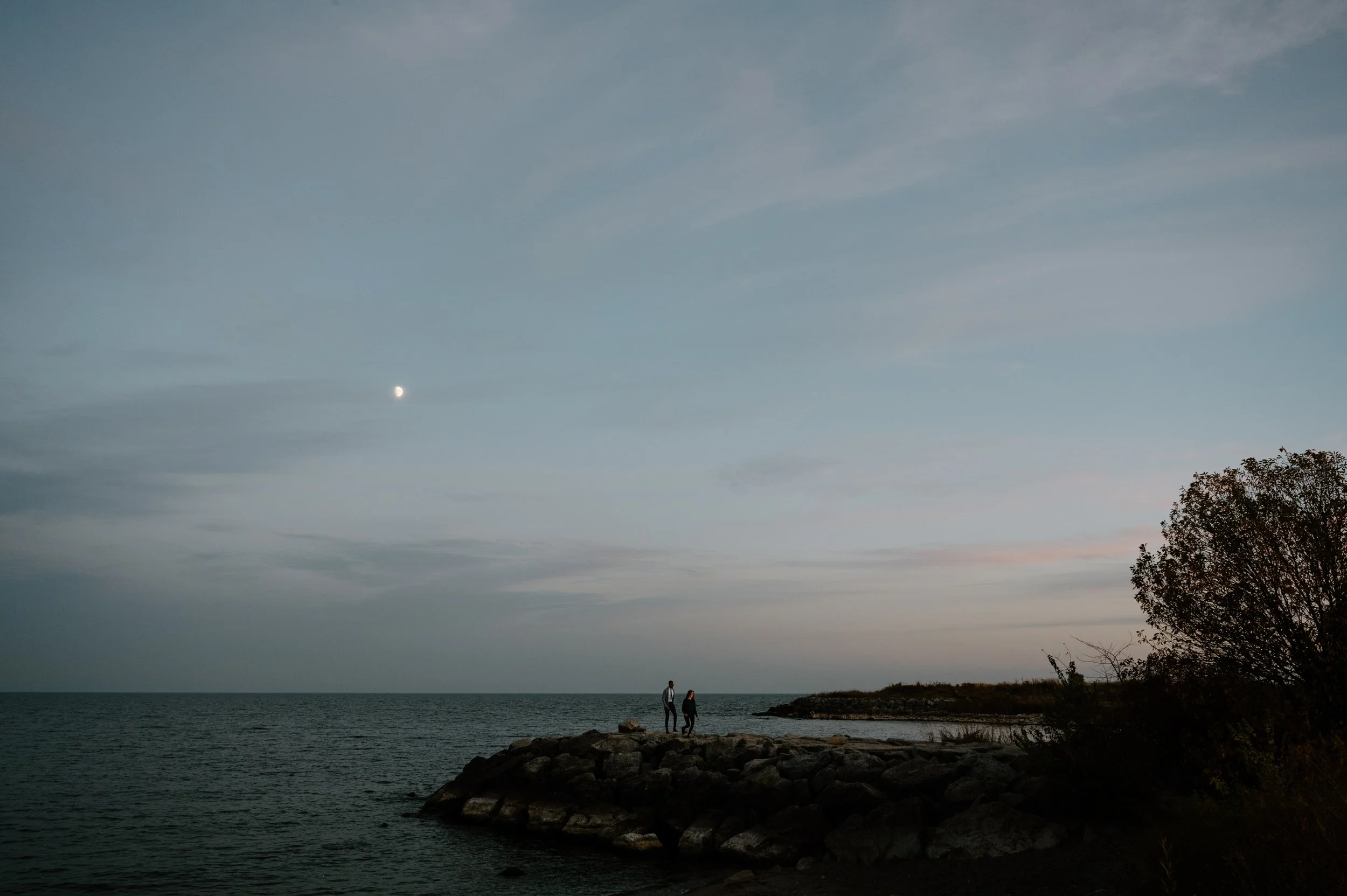 Wide landscape photo of an engaged couple walking along the waterfront at sunset, a setting that evokes both Toronto and Vancouver