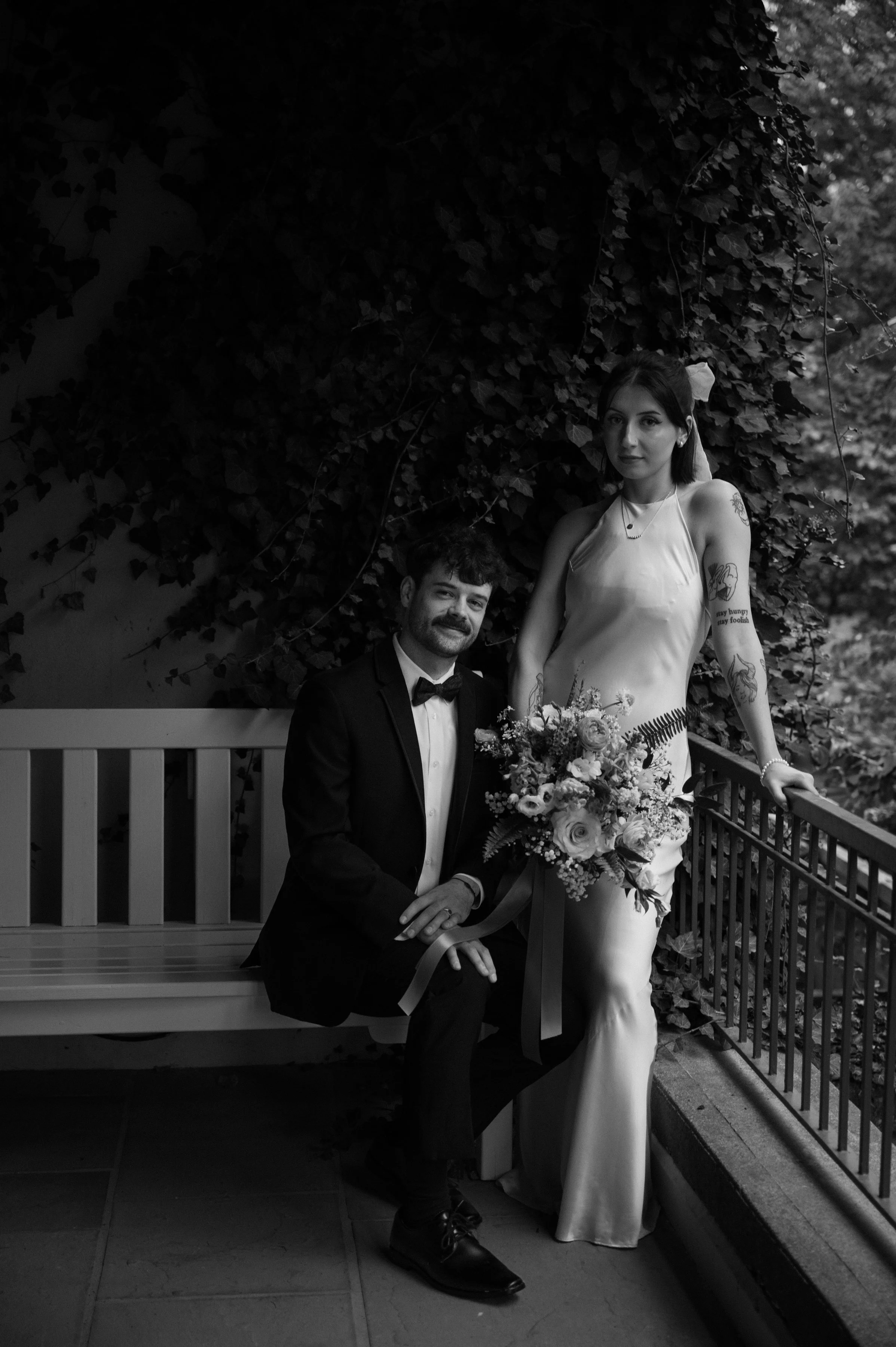 Black and white portrait of a bride and groom seated on a bench in front of a vine-covered garden wall at Langdon Hall