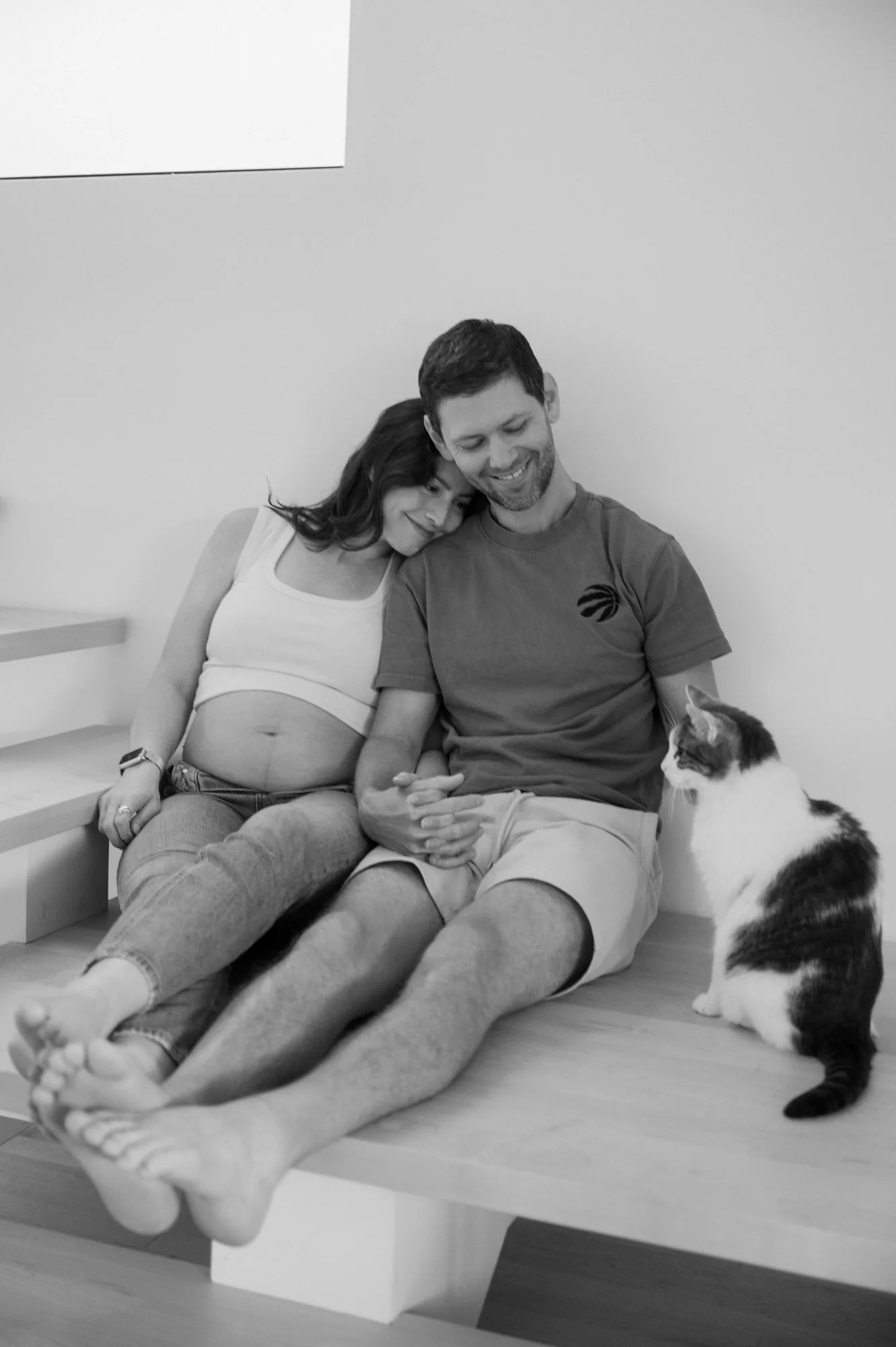 Black and white photo of a couple sitting on their stairway landing with their cat during an at-home maternity session in Toronto
