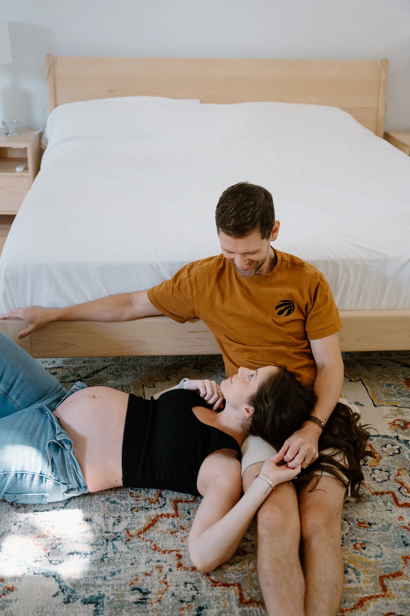 Documentary photo of a couple sitting at the foot of their bed, mom to be resting on her partner's lap during an at-home maternity session in Toronto