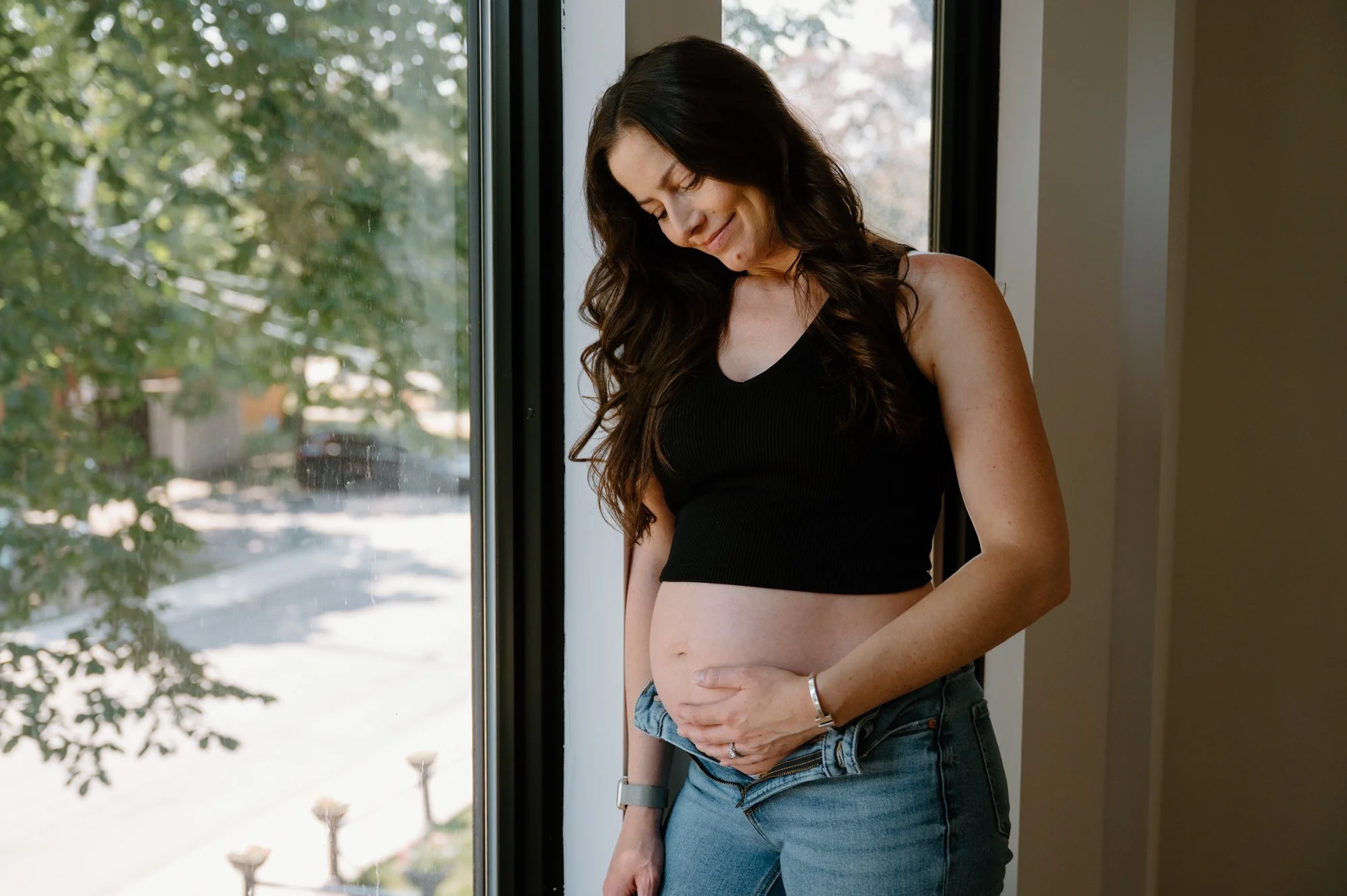 Candid emotional photo of a mom to be standing by a window embracing her belly during an at-home maternity session in Toronto