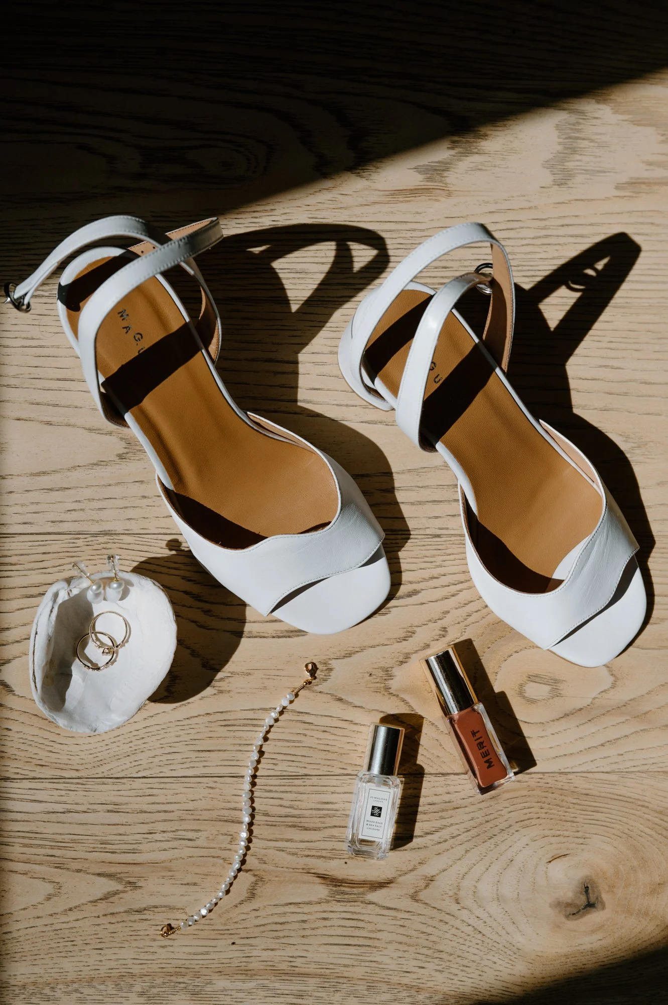 Flat lay of bridal details including Maguire shoes, Bluboho rings, perfume, lipgloss, and a sea shell at a destination wedding in Newfoundland