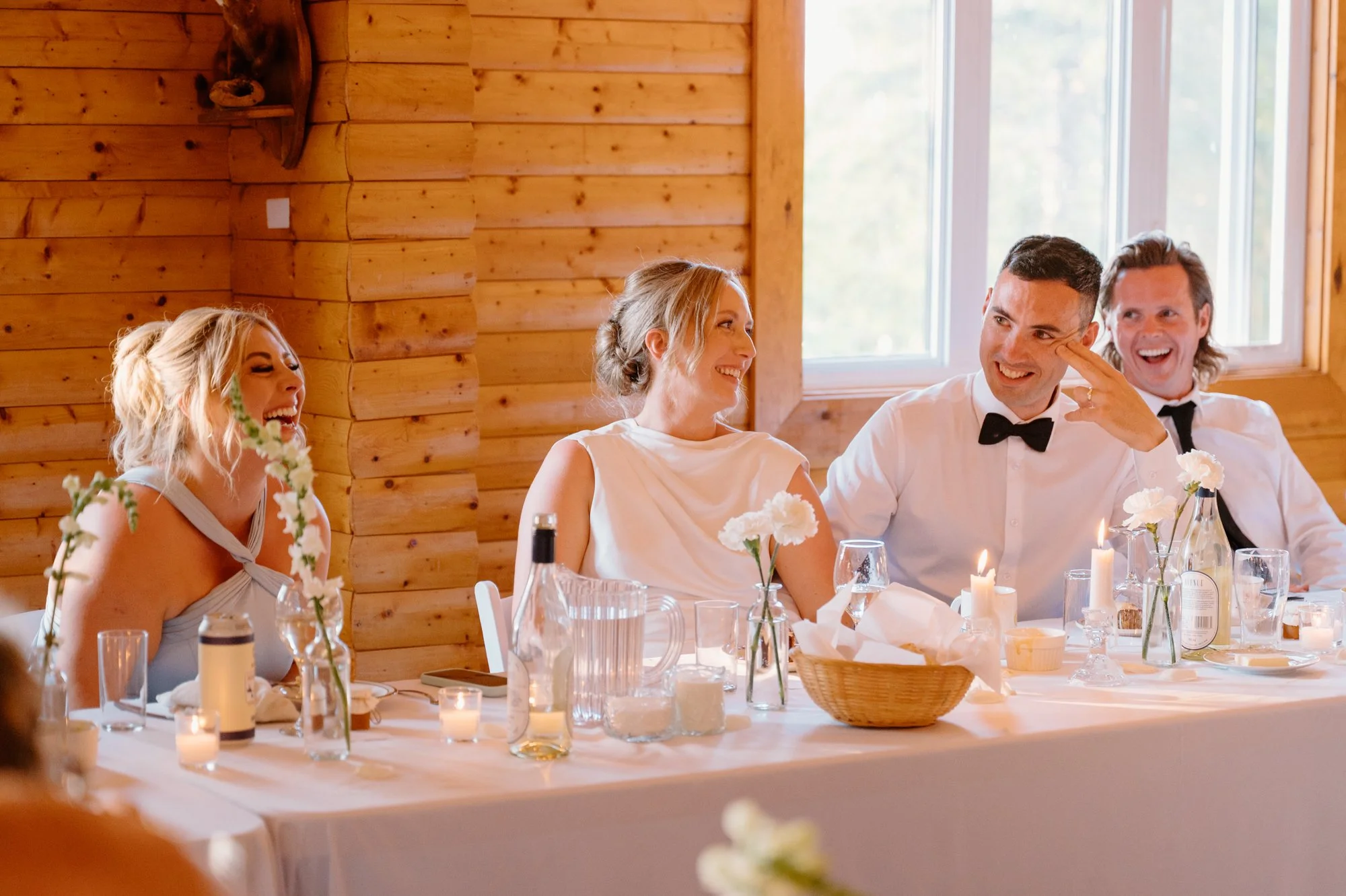 Candid photo of a bride and groom laughing together while a guest delivers a speech at their destination wedding reception
