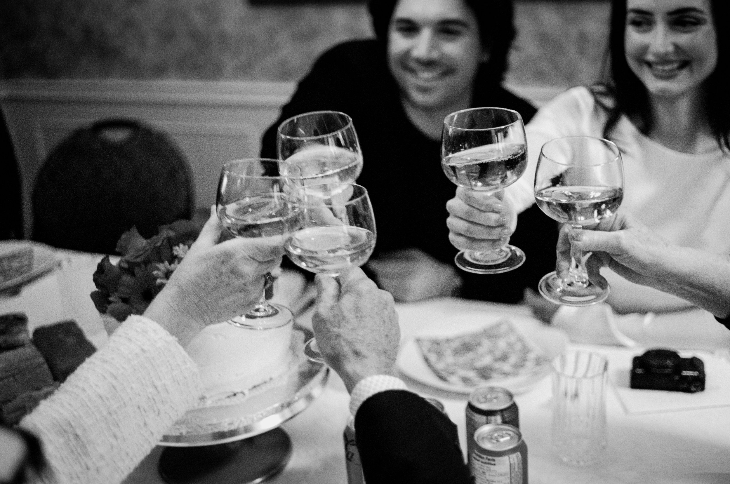 Black and white candid film photograph of wedding guests raising champagne glasses in a cheers during an intimate at-home wedding dinner in Toronto