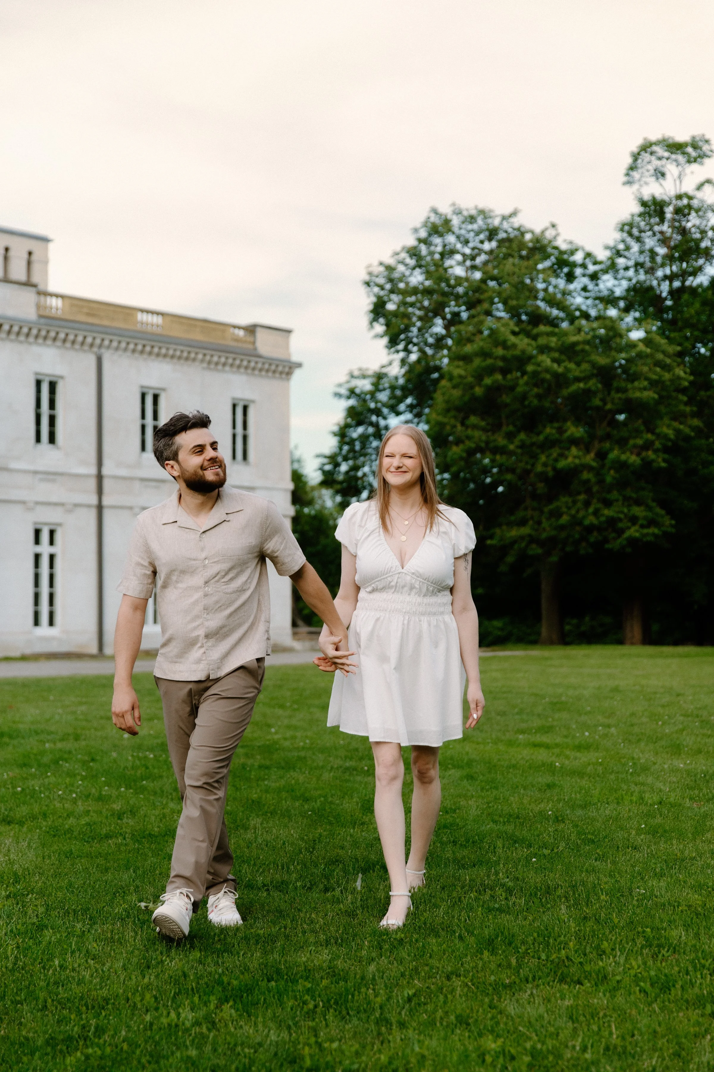 Candid photo of a couple walking and smiling on the front lawn of Dundurn Castle during their engagement session, dressed in a complementary neutral colour palette