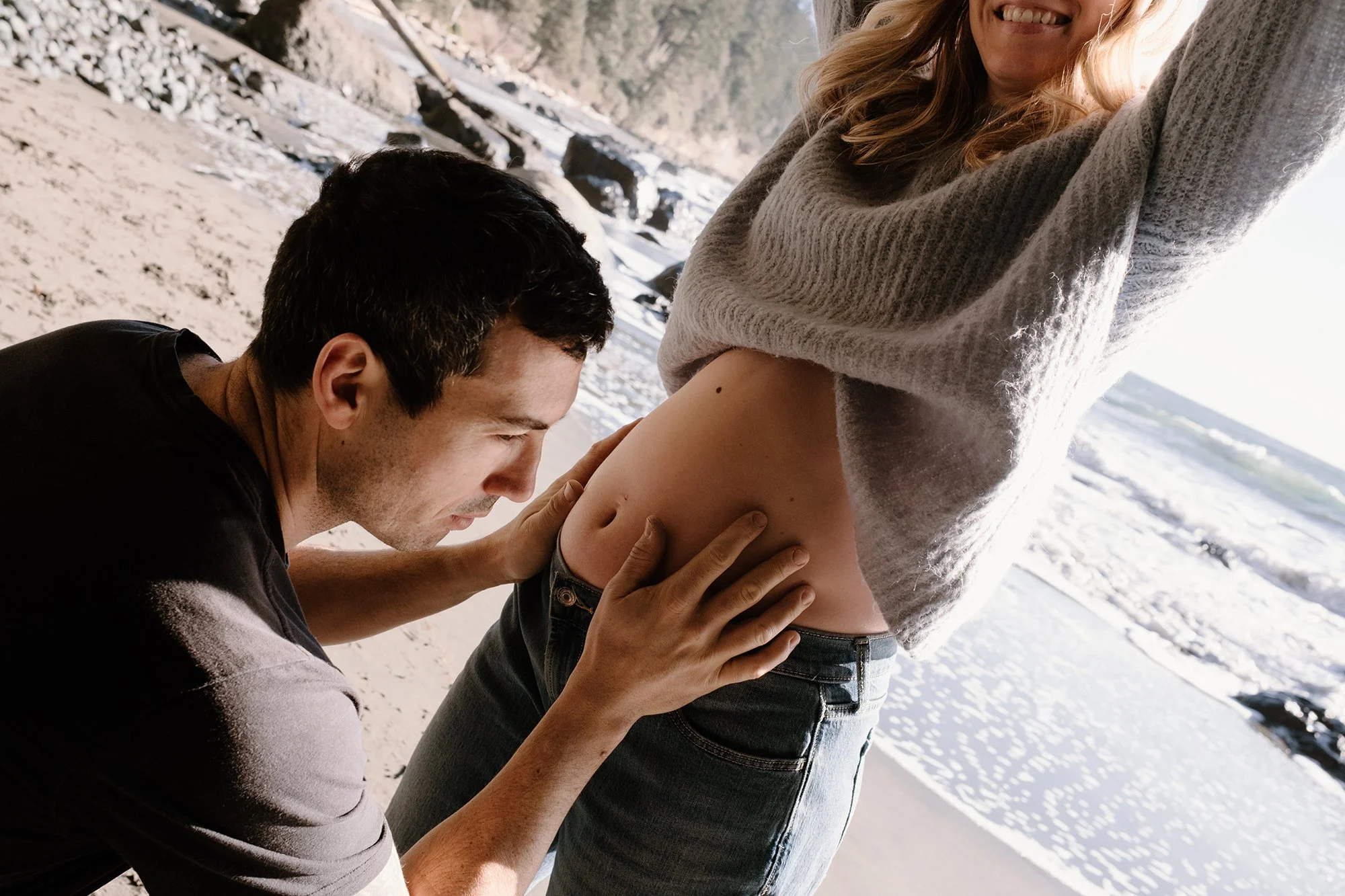Editorial maternity portrait of a couple standing at the waterfront in Victoria, BC as he holds her pregnancy bump and she smiles