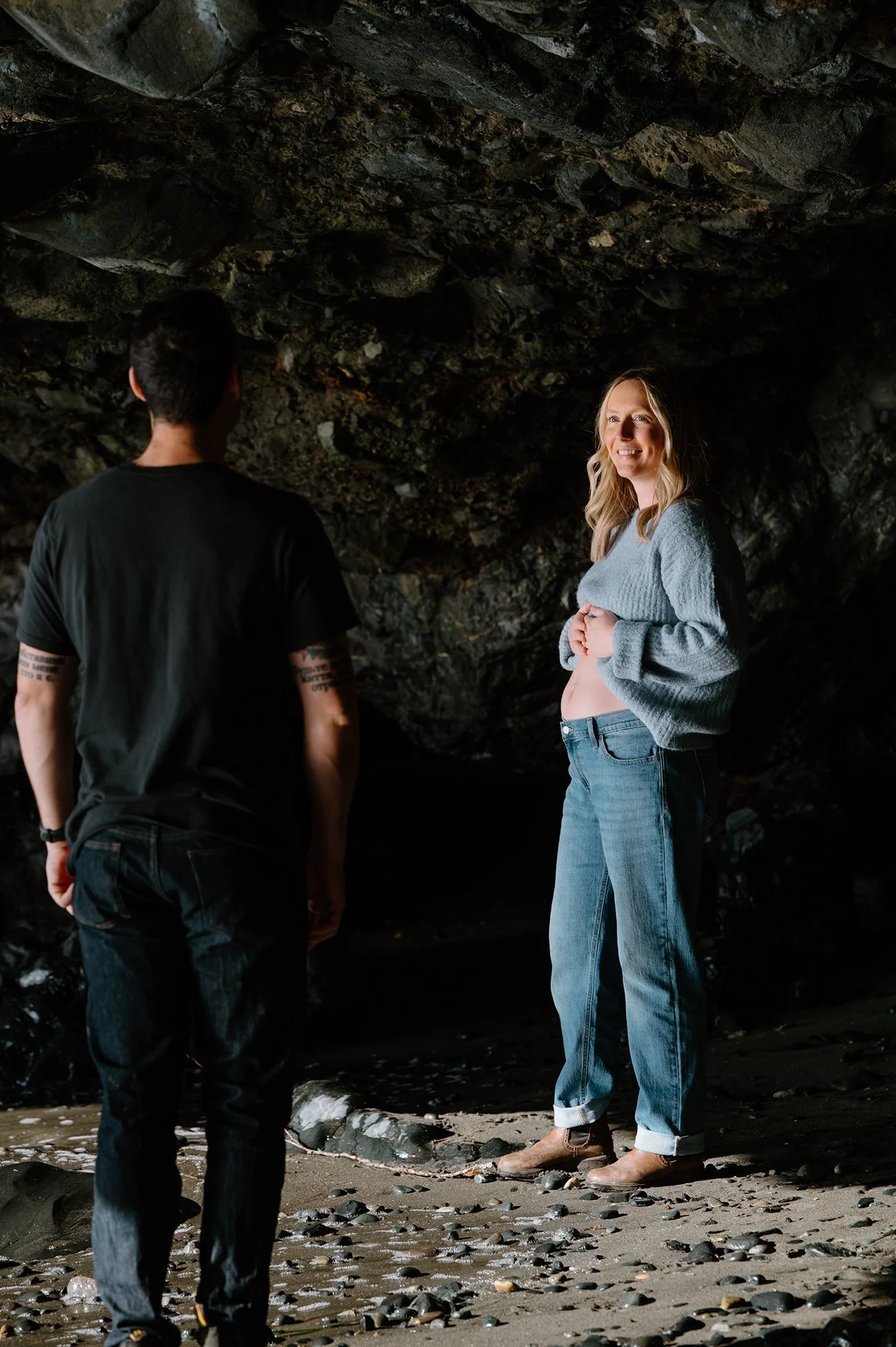 Maternity photo of a couple standing inside the cave at Mystic Beach on Vancouver Island, she faces out towards the ocean as he stands facing her