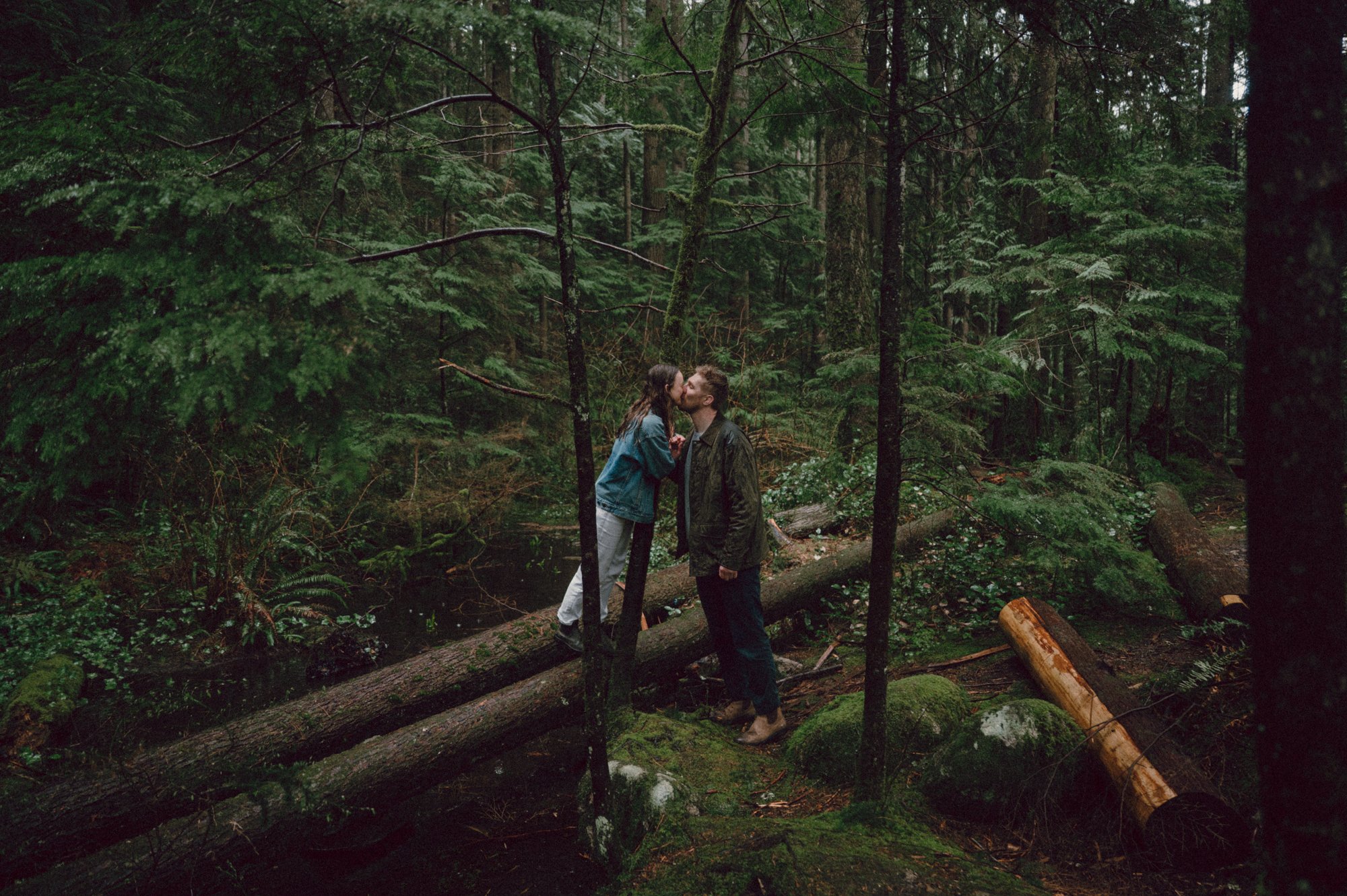 Candid photo of a couple kissing as they walk through the forest in Lynn Valley, she is standing on logs as he helps her along