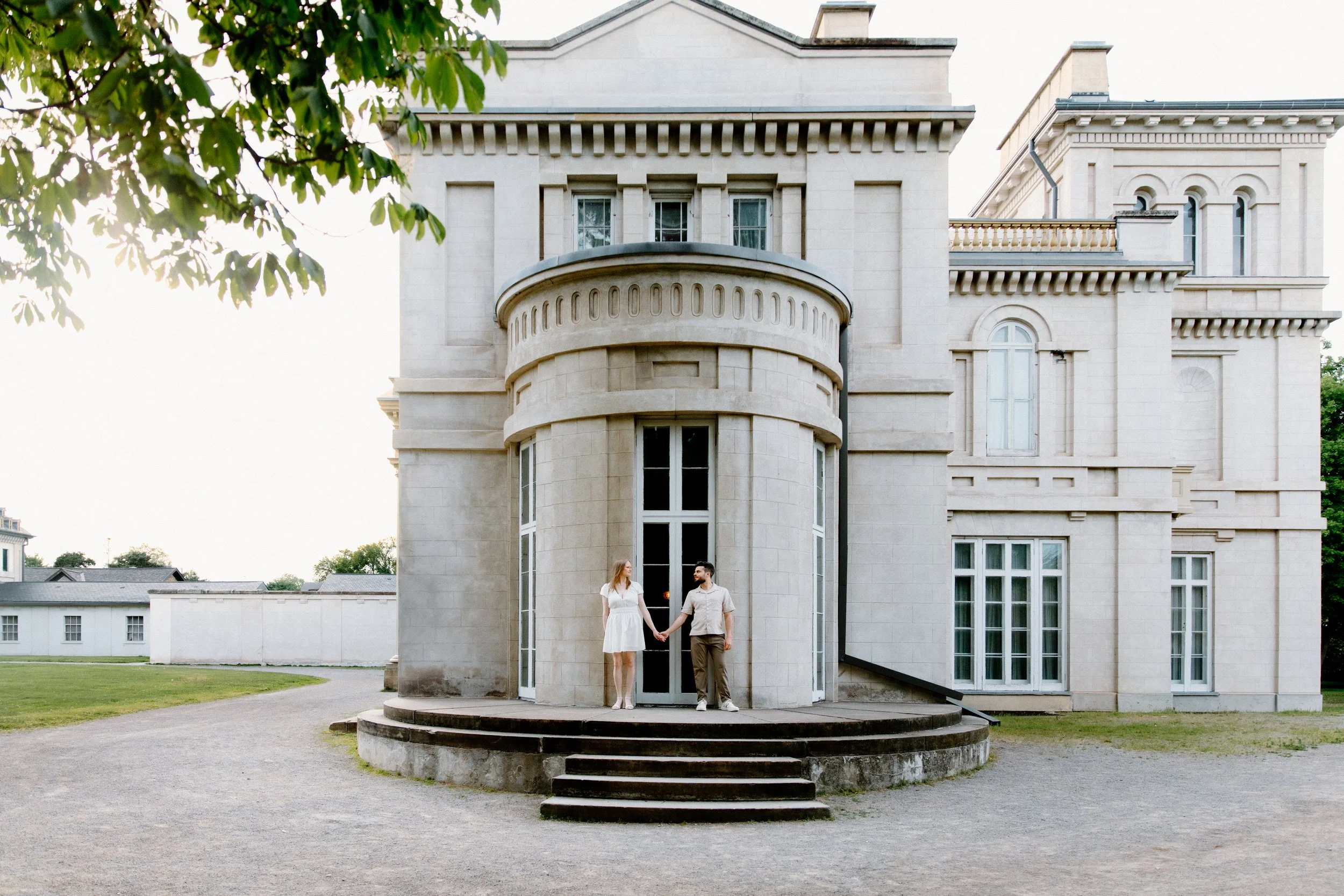 Wide photo of a couple standing at the side of Dundurn Castle with the sunset behind them during their engagement session