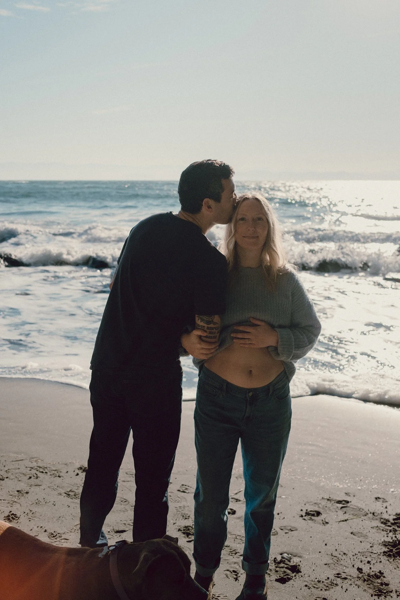 Film photograph of a couple standing at the ocean on Vancouver Island, their dog slightly in frame, as he kisses her head while she shows her pregnancy bump