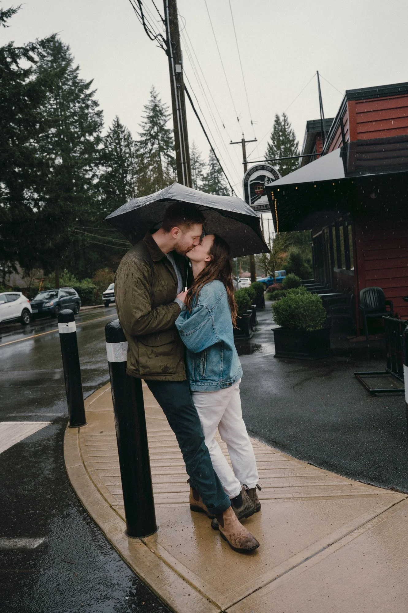Romantic engagement photo of a couple kissing under an umbrella near the End of the Line General Store in Lynn Valley, Vancouver on a rainy day