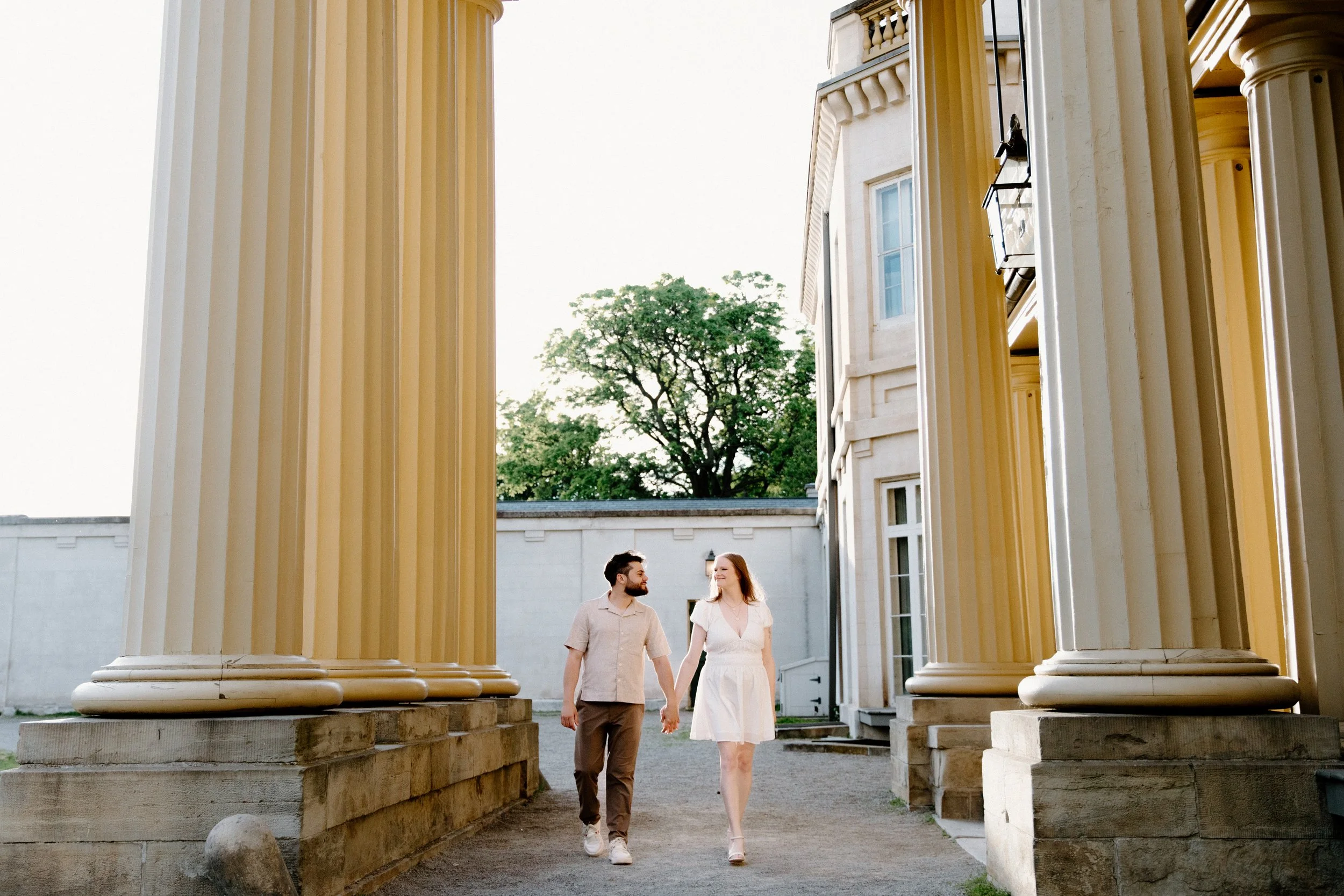 Candid photo of a couple walking between the pillars at the front of Dundurn Castle with the sunset behind them during their engagement session