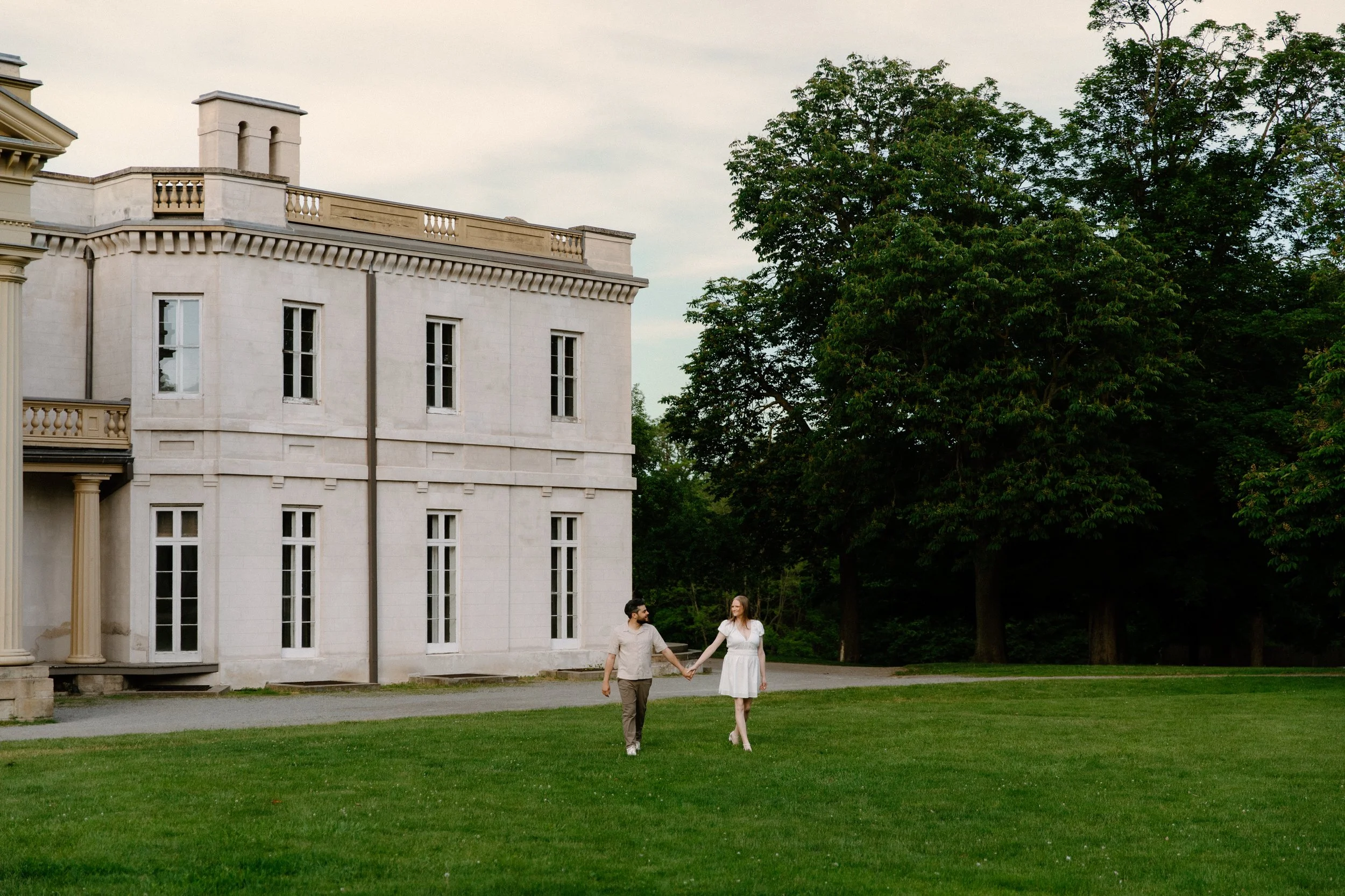 Wide photo of a couple walking on the front lawn of Dundurn Castle, architecture of the castle prominent in the background