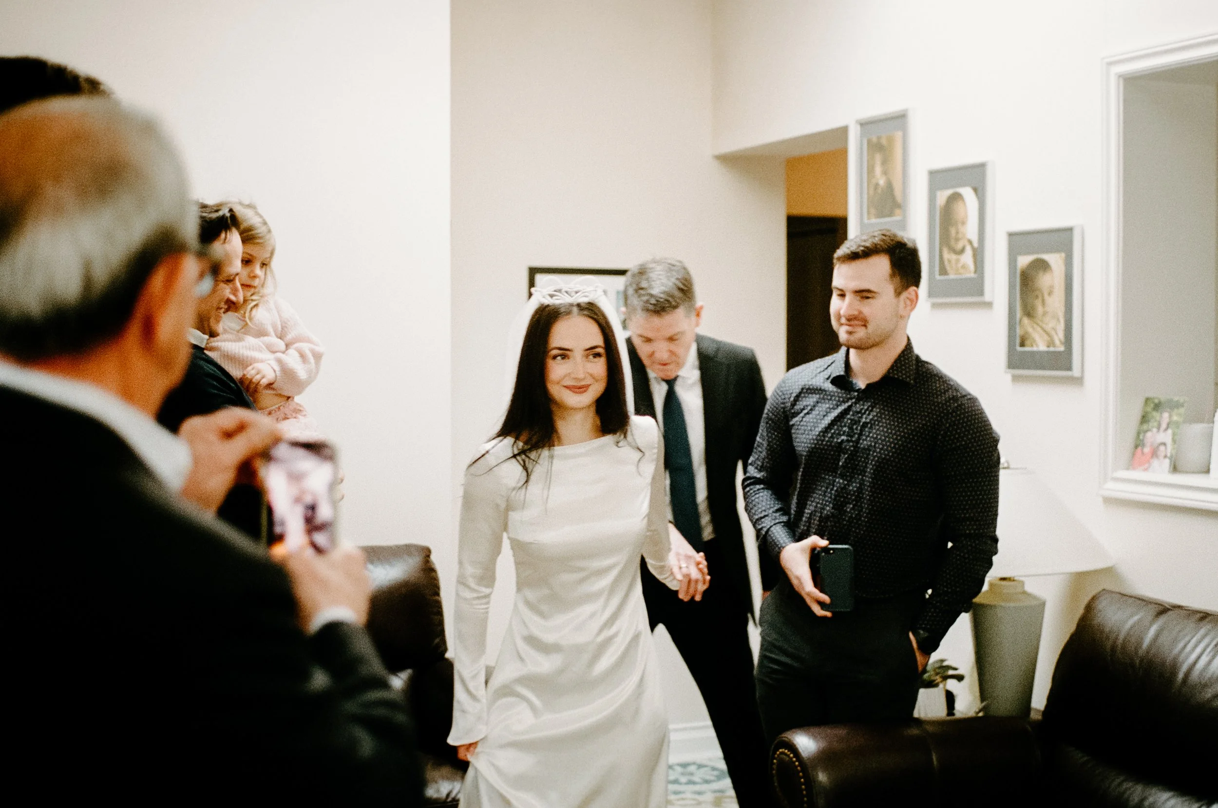 Candid film photograph of a bride walking into her living room ceremony with her father as guests smile and clap around them