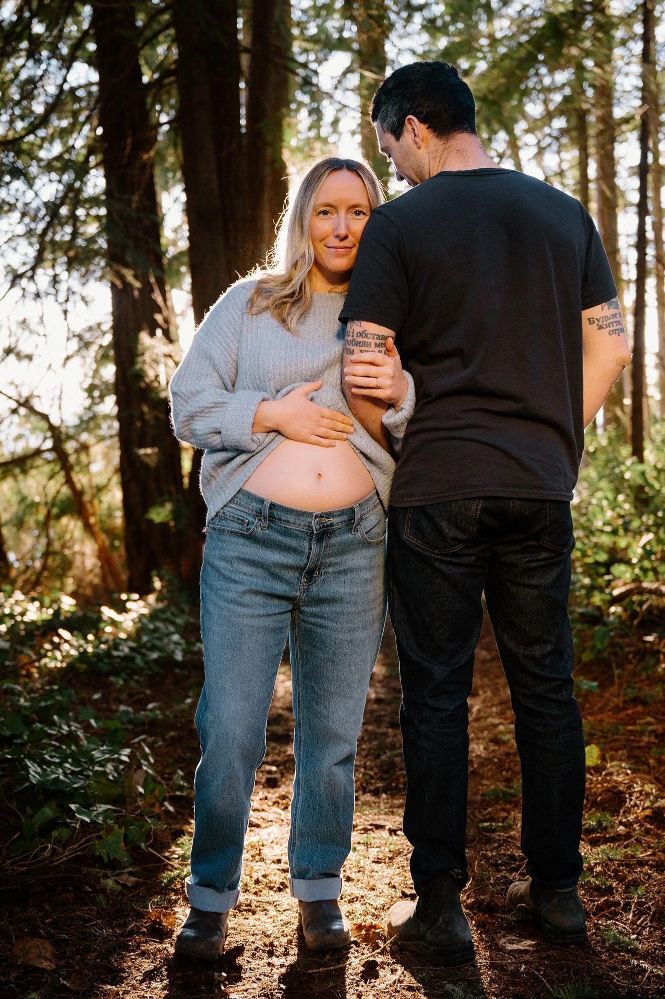 Maternity photo of a couple standing side by side on a trail at sunset on Vancouver Island, she faces the camera with her bump showing as he looks away