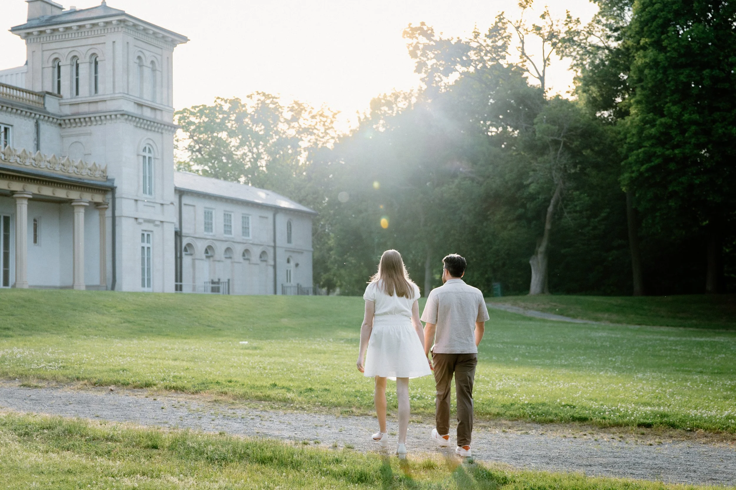 Romantic photo of a couple walking towards Dundurn Castle as the sun sets behind them during their engagement session