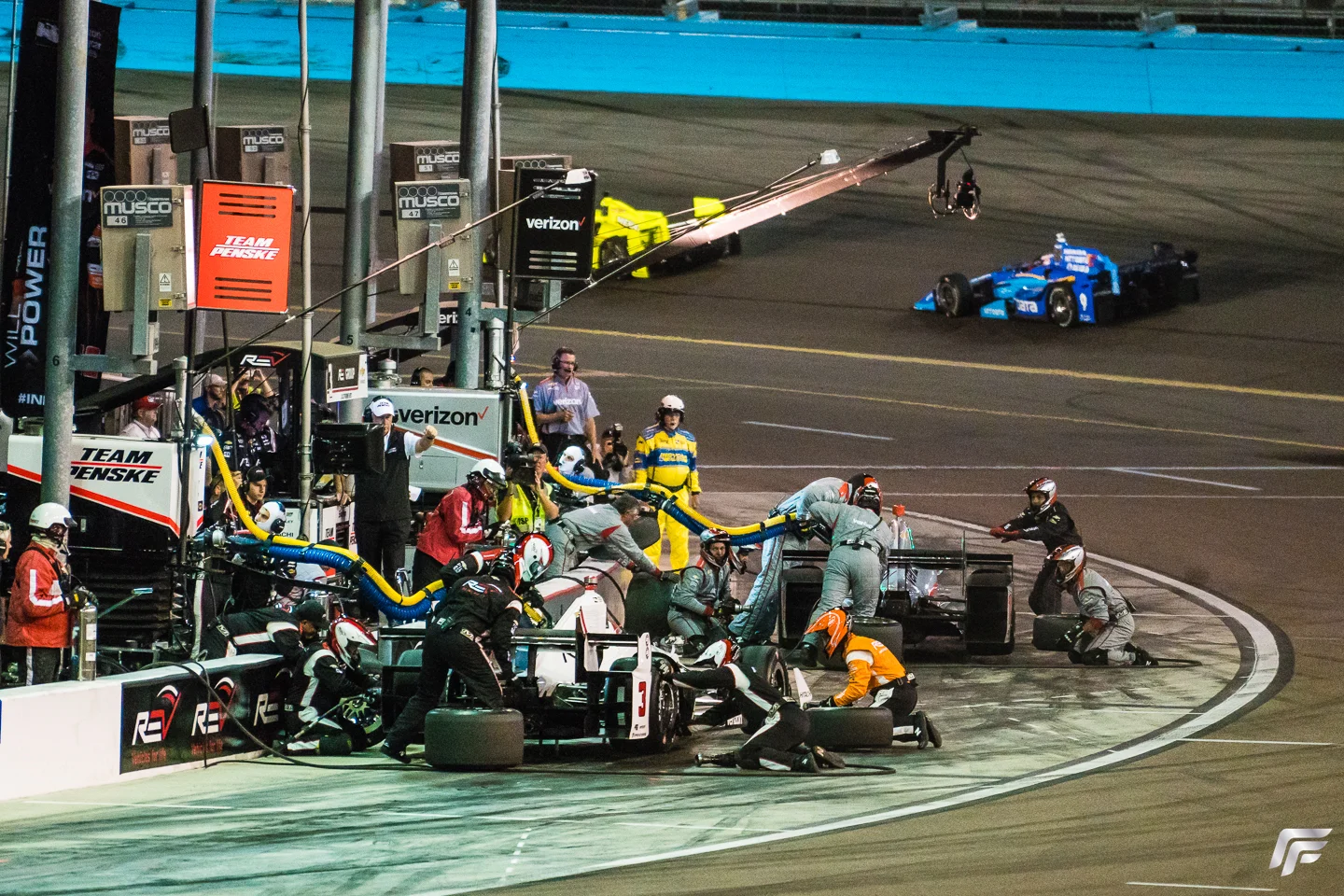 Simon Pagenaud’s brightly colored Menards Honda can be seen in the distance remaining on course while Castoneves and Power are in the pits.