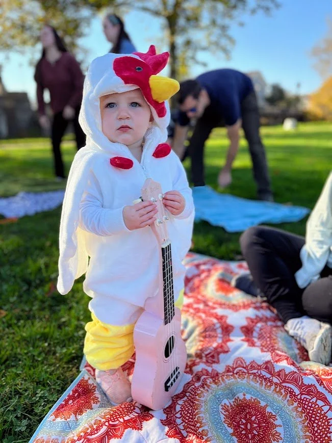 Babies and toddlers dancing and playing instruments at an outdoor music and movement playgroup in Kerr Park in costume at Halloween