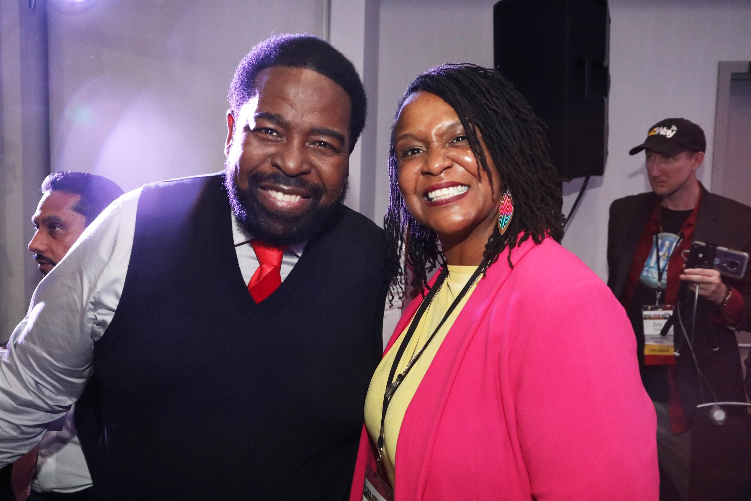 A man and woman smiling at the camera. The man has a beard, dark hair, and is wearing a white shirt with a black vest and red tie. The woman has braided hair, large colorful earrings, a yellow top, and a bright pink jacket. In the background, two other men are visible, one wearing a cap and holding a phone.