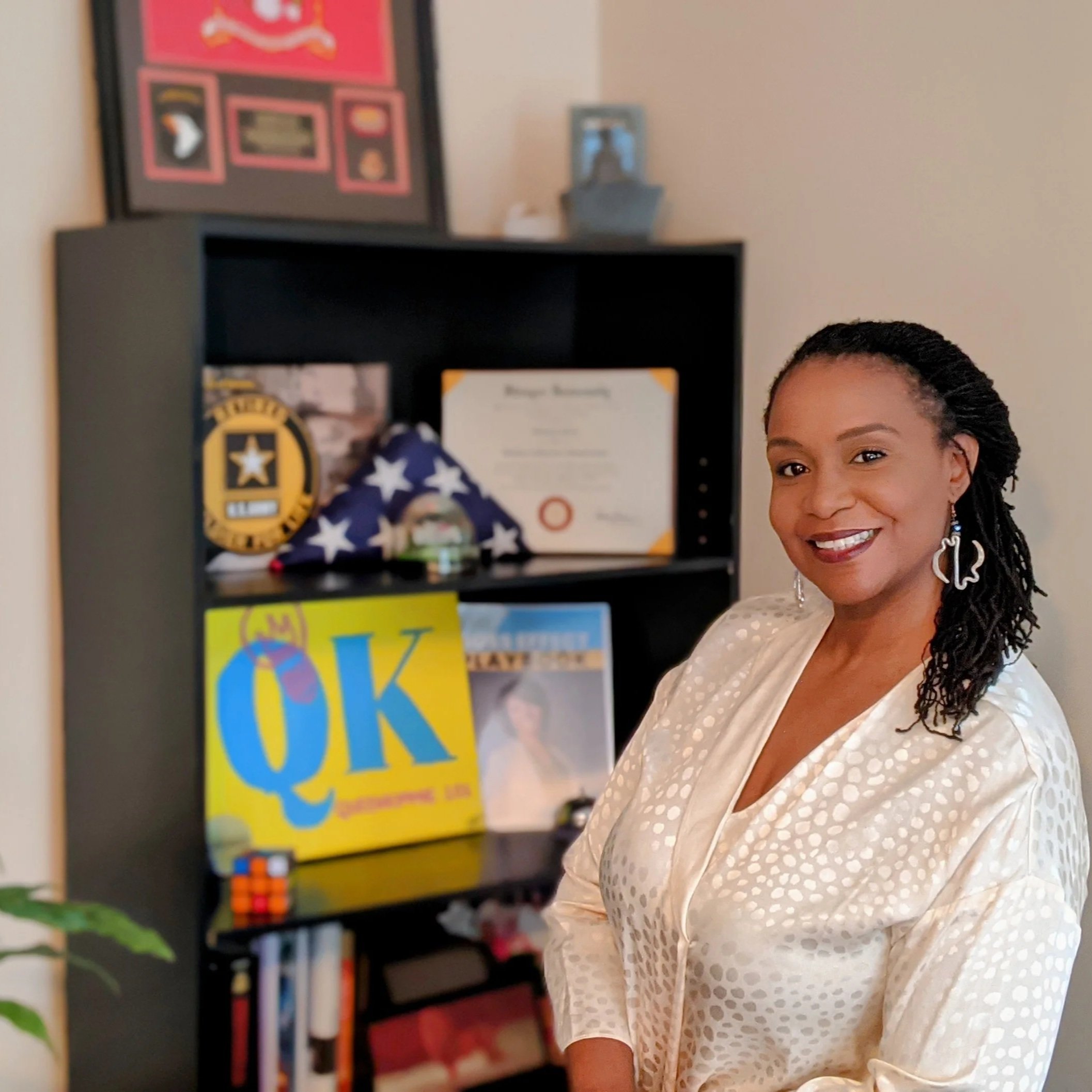 A smiling woman with braided hair wearing earrings and a cream-colored blouse standing in front of a bookshelf with awards, a folded American flag, and framed documents, including a colorful sign with 'QK' on it.