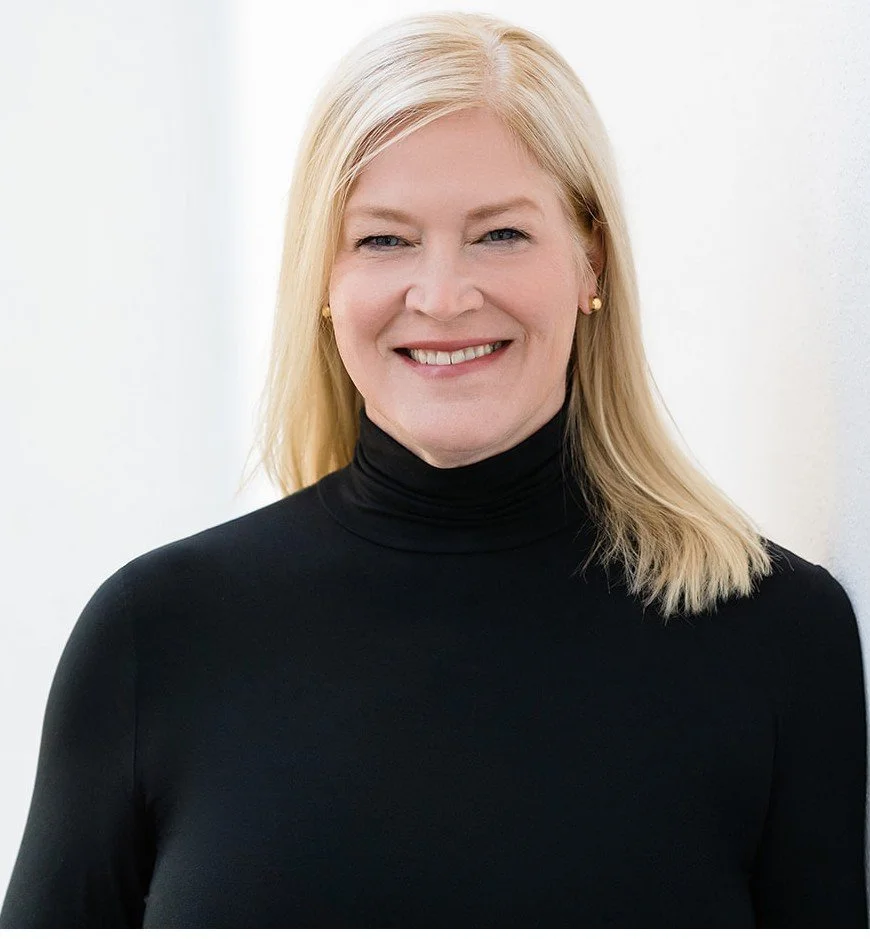 A blonde woman smiling, wearing a black turtleneck and small earrings, standing against a plain white background.
