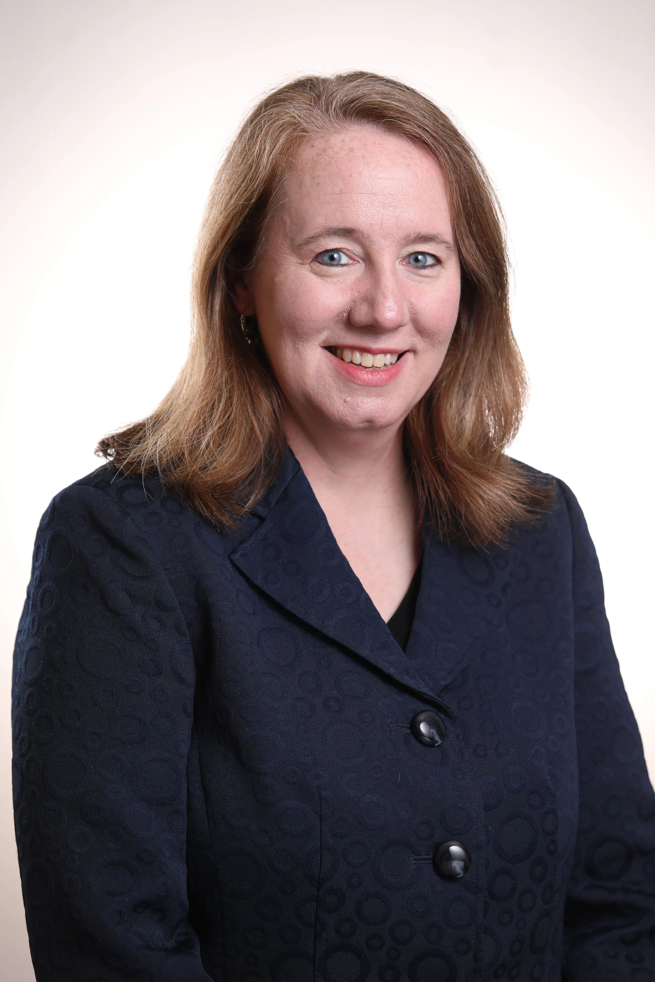 Headshot of a woman with reddish-brown hair, blue eyes, smiling, wearing a dark navy blazer with large black buttons, against a plain white background.