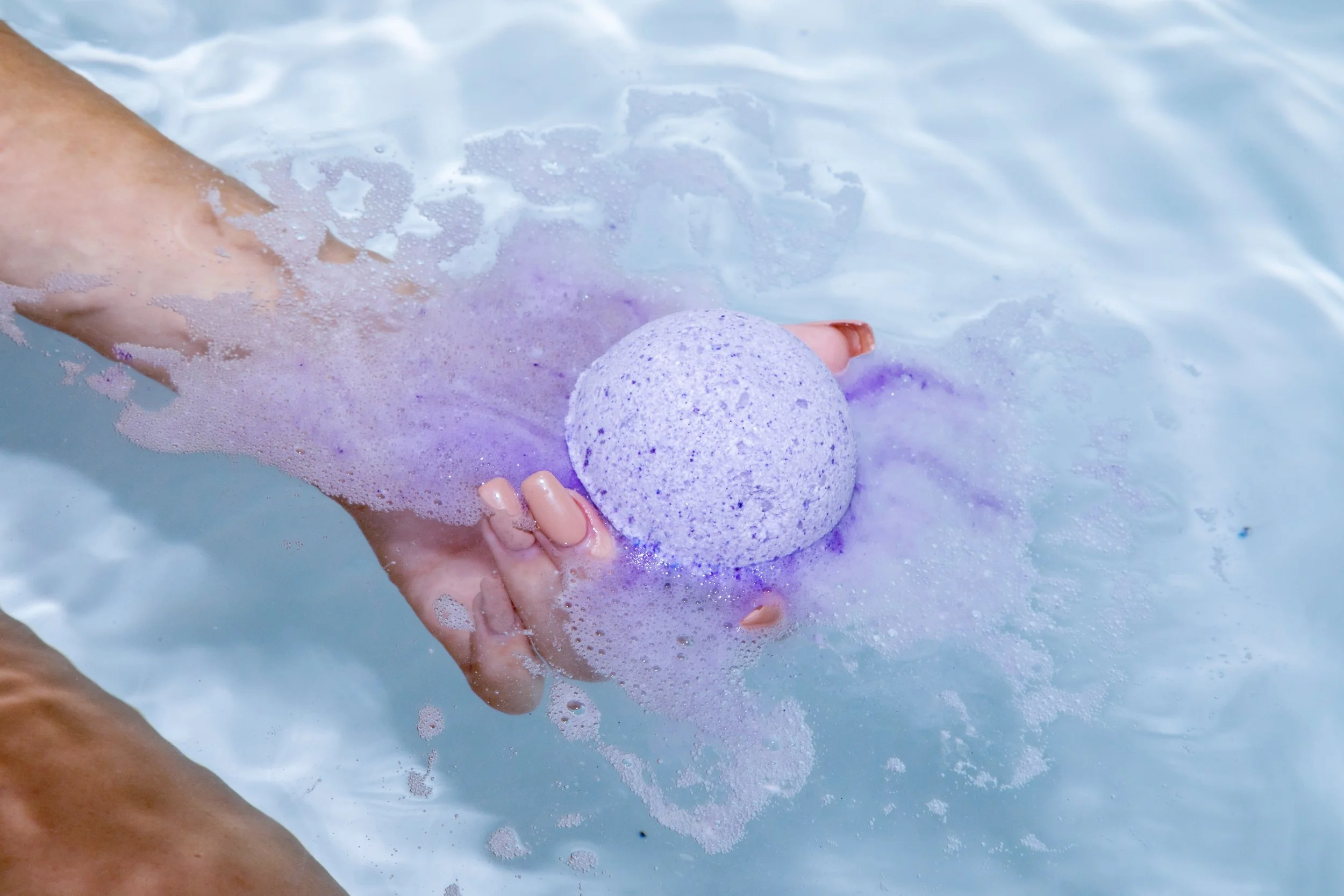 Hand holding purple bath bomb that is fizzing in water.