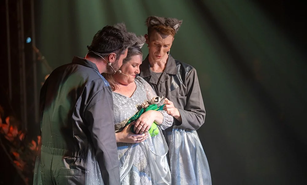 Three stage actors, the middle actor holding a small animal like a baby, in costume in a theatre production called Where the Wind Blew.