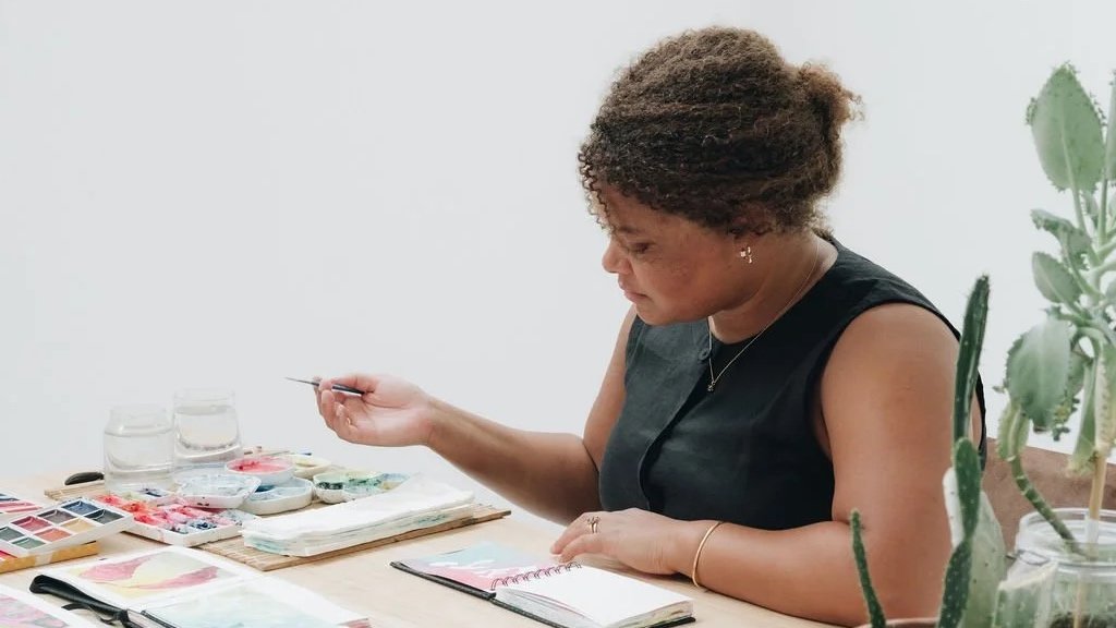 A woman with curly hair, wearing a black sleeveless top, sitting at a table with watercolor paints, paintbrushes, notebooks, and two glasses of water, in a room with white walls and a green potted cactus plant.
