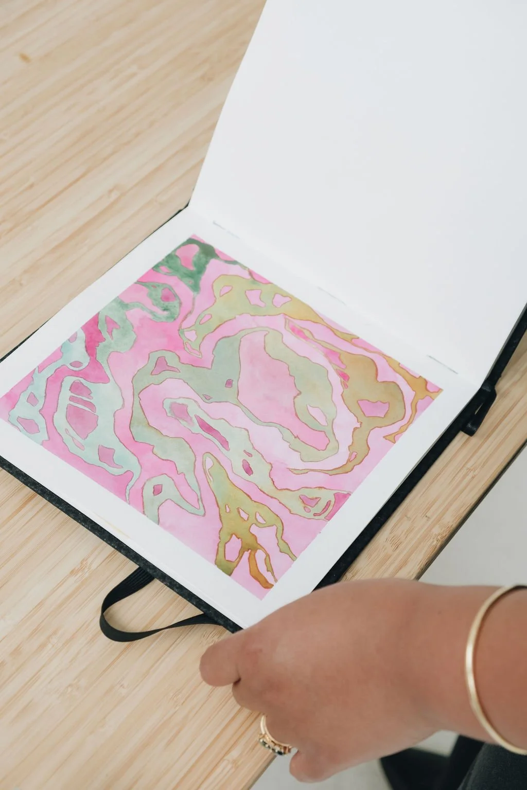 Open sketchbook showing abstract watercolor painting with pink, green, and gold colors, placed on a wooden table, with a person's hand and jewelry visible.
