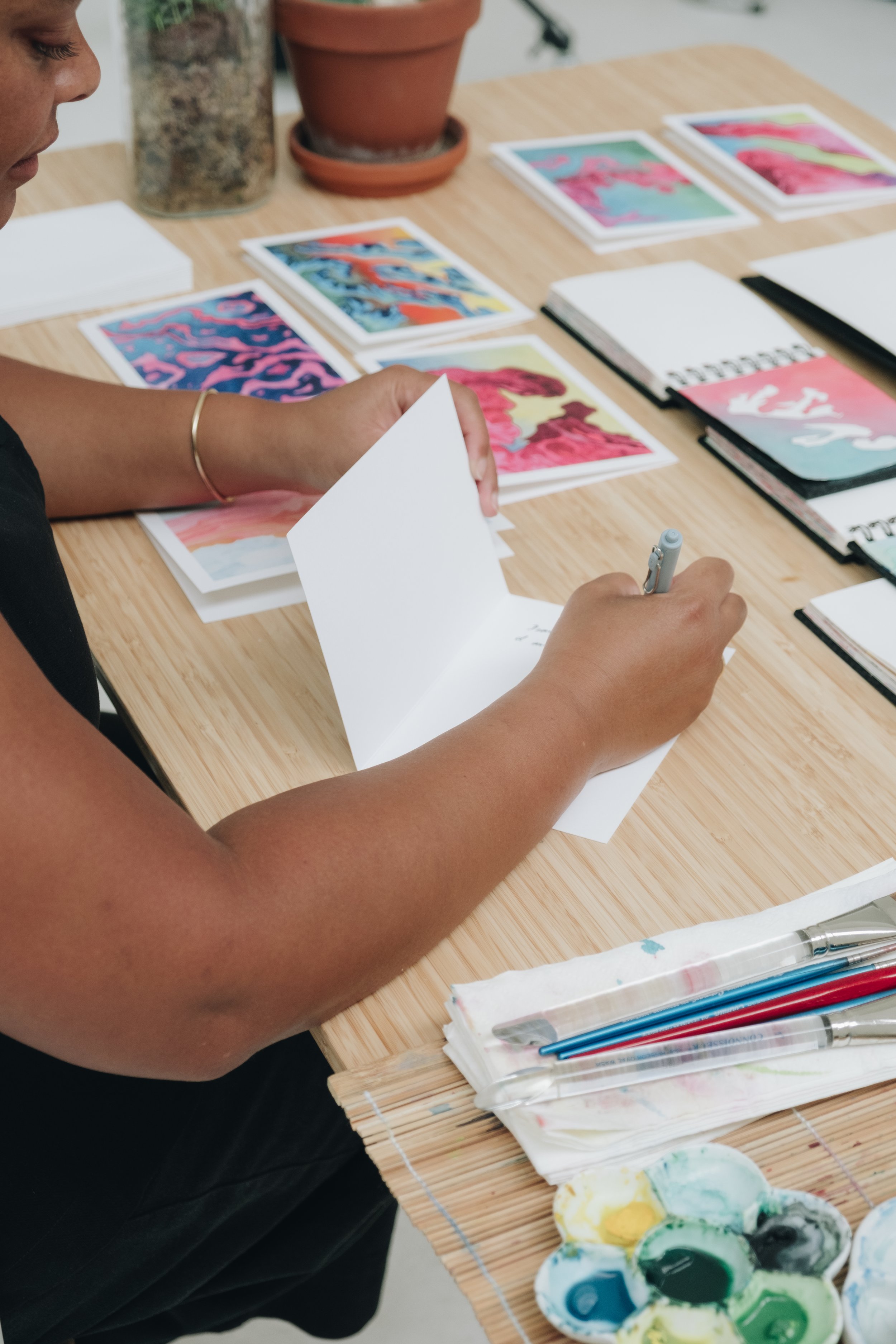 A person sitting at a wooden table reviewing colorful abstract art prints, with notebooks, paint brushes, and watercolor palettes nearby.
