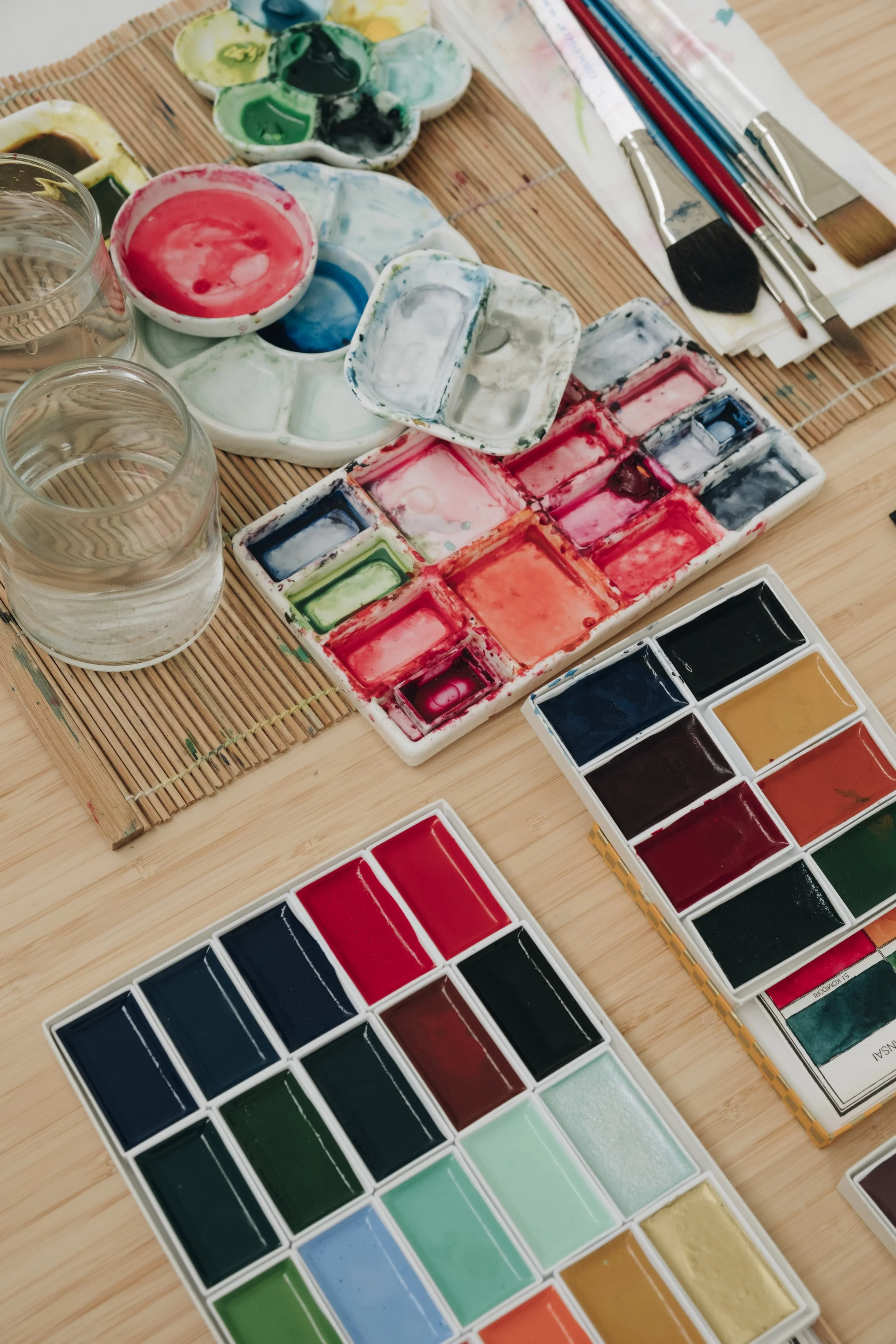 Art supplies on a wooden table, including watercolor paint palettes, jars of water, and paintbrushes.