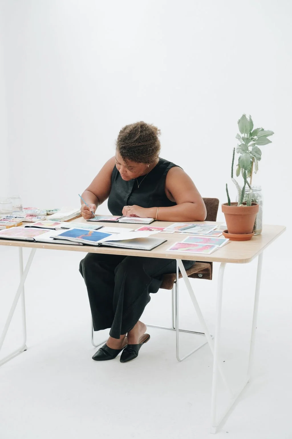 A woman sitting at a light wood table, working on art projects with various papers and watercolor paints, in a minimal white space with a potted plant nearby.