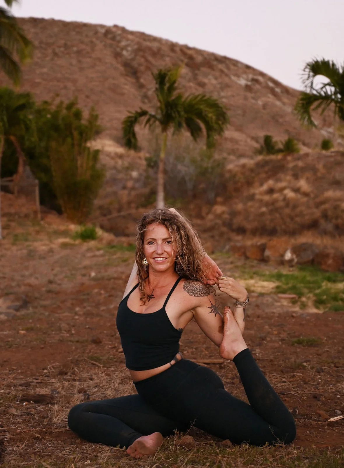A woman practicing yoga outdoors on a dirt ground with palm trees and hilly terrain in the background during sunset.